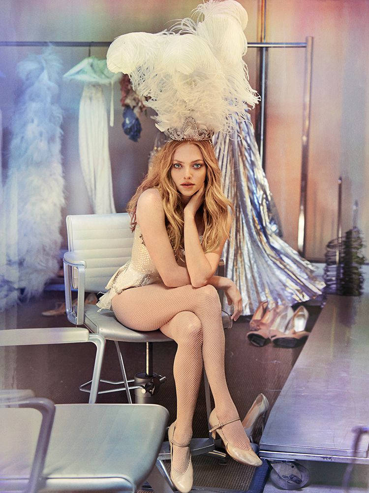 Amanda Seyfried 桌布 壁紙 — 一位金髮女性戴著高聳的白色羽毛頭飾和漁網襪，坐在更衣室的椅子上。