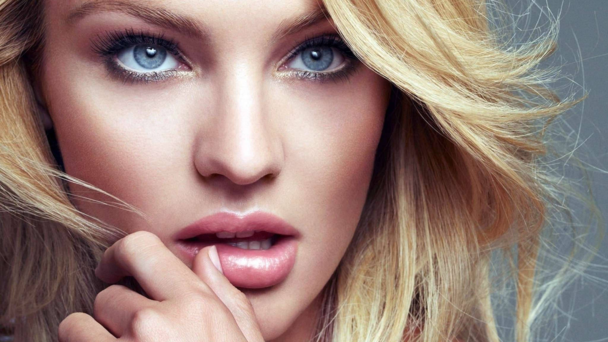 Candice 桌布 壁紙 — 一位金髮女性的特寫肖像，擁有銳利的藍色眼睛和光澤的嘴唇，她的手輕輕觸碰嘴巴，背景為柔和的灰色。