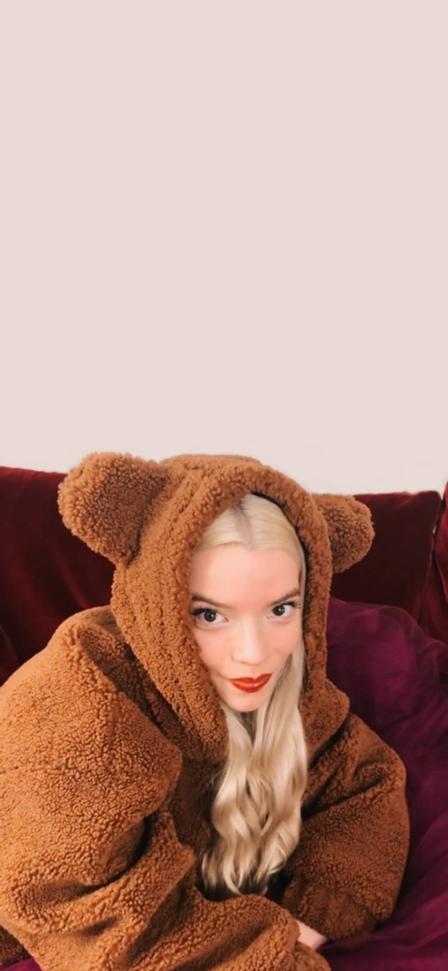 Anya 桌布 壁紙 — 一位金髮女性穿著棕色泰迪熊連體衣，在柔軟的紅色沙發上休息，背景柔和中性。
