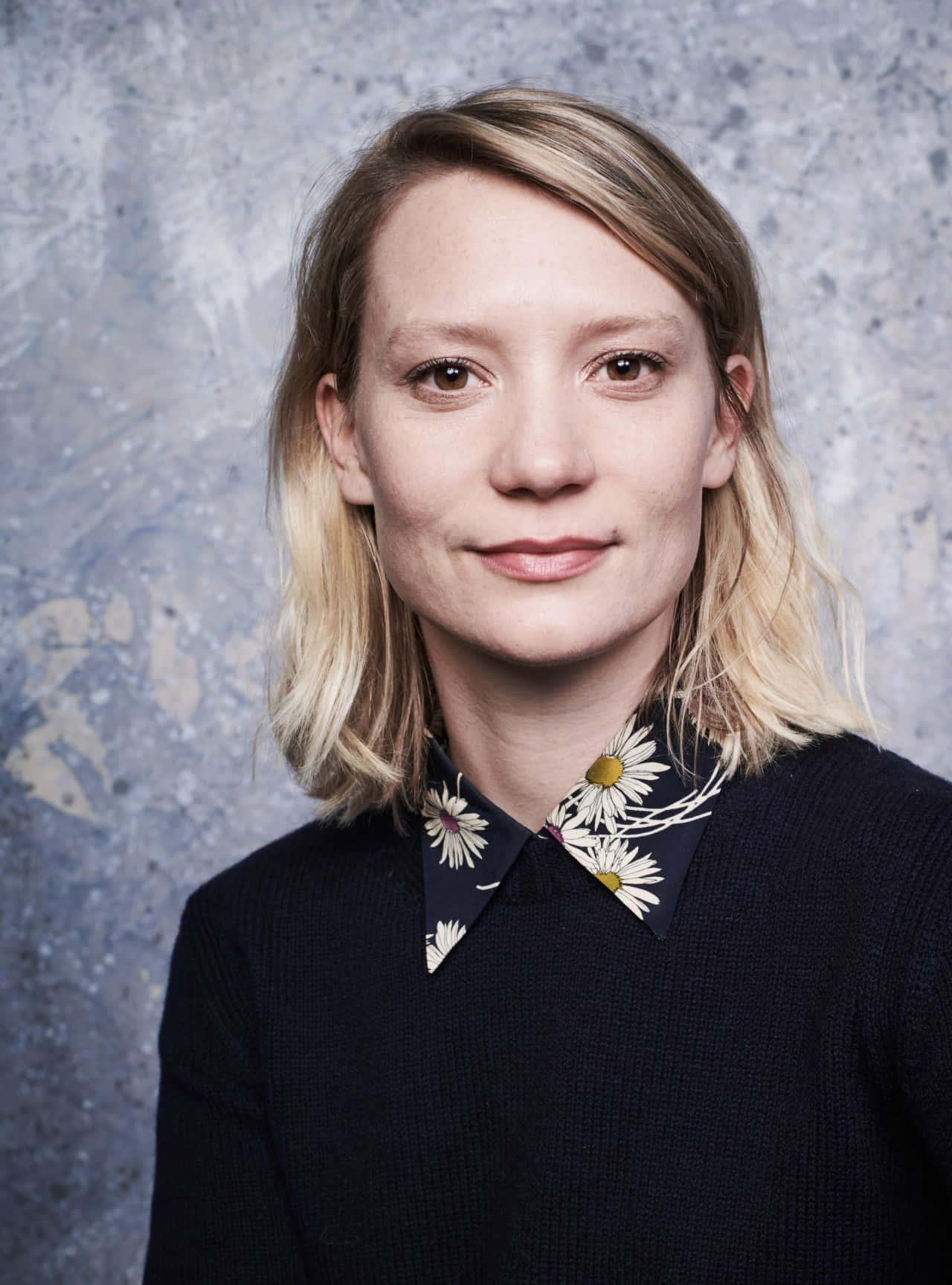 Mia Wasikowska 桌布 壁紙 — 一位金髮女性穿著深色毛衣，衣領帶有雛菊圖案，背景是帶紋理的灰藍色牆壁。