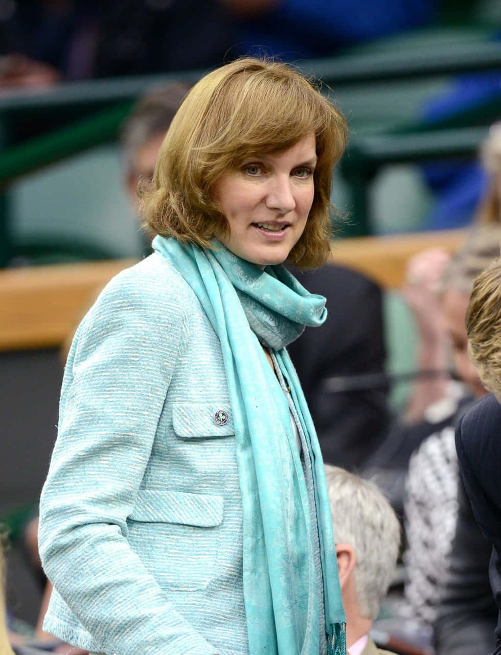 Fiona Bruce 桌布 壁紙 — 一位金髮女性穿著淺藍色質感外套和相配的圍巾，站在體育場的人群中。