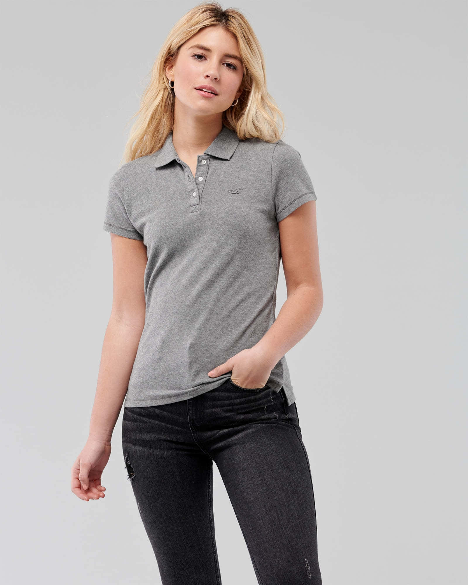 Hollister 桌布 壁紙 — 一位金髮女性穿著灰色Polo衫和黑色牛仔褲，背景為純灰色。