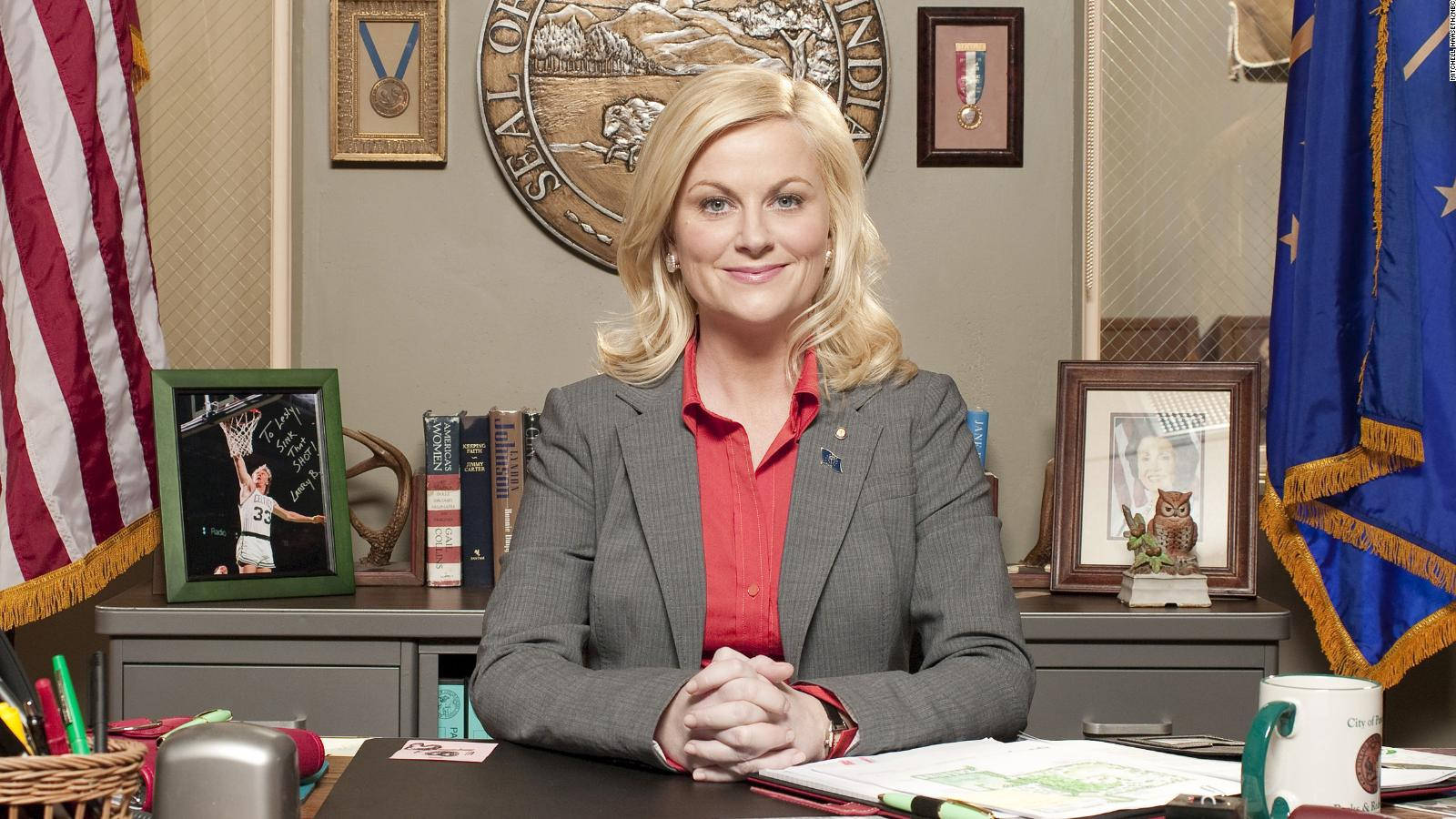 parks and recreation 桌布 壁紙 — 一位金髮女性穿著灰色西裝和紅色襯衫，自信地對著鏡頭微笑，坐在一張雜亂的辦公桌前，桌上裝飾著美國國旗和裱框照片。