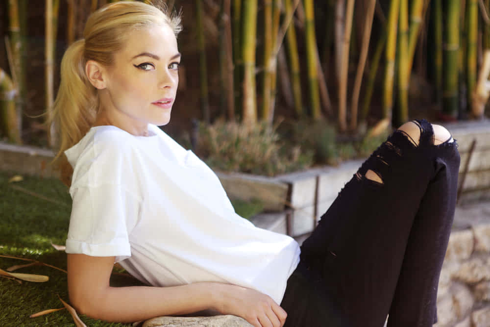 Jaime King 桌布 壁紙 — 一位金髮女性穿著白色T恤和破洞黑色牛仔褲，在身後有竹稈的草地上休息。