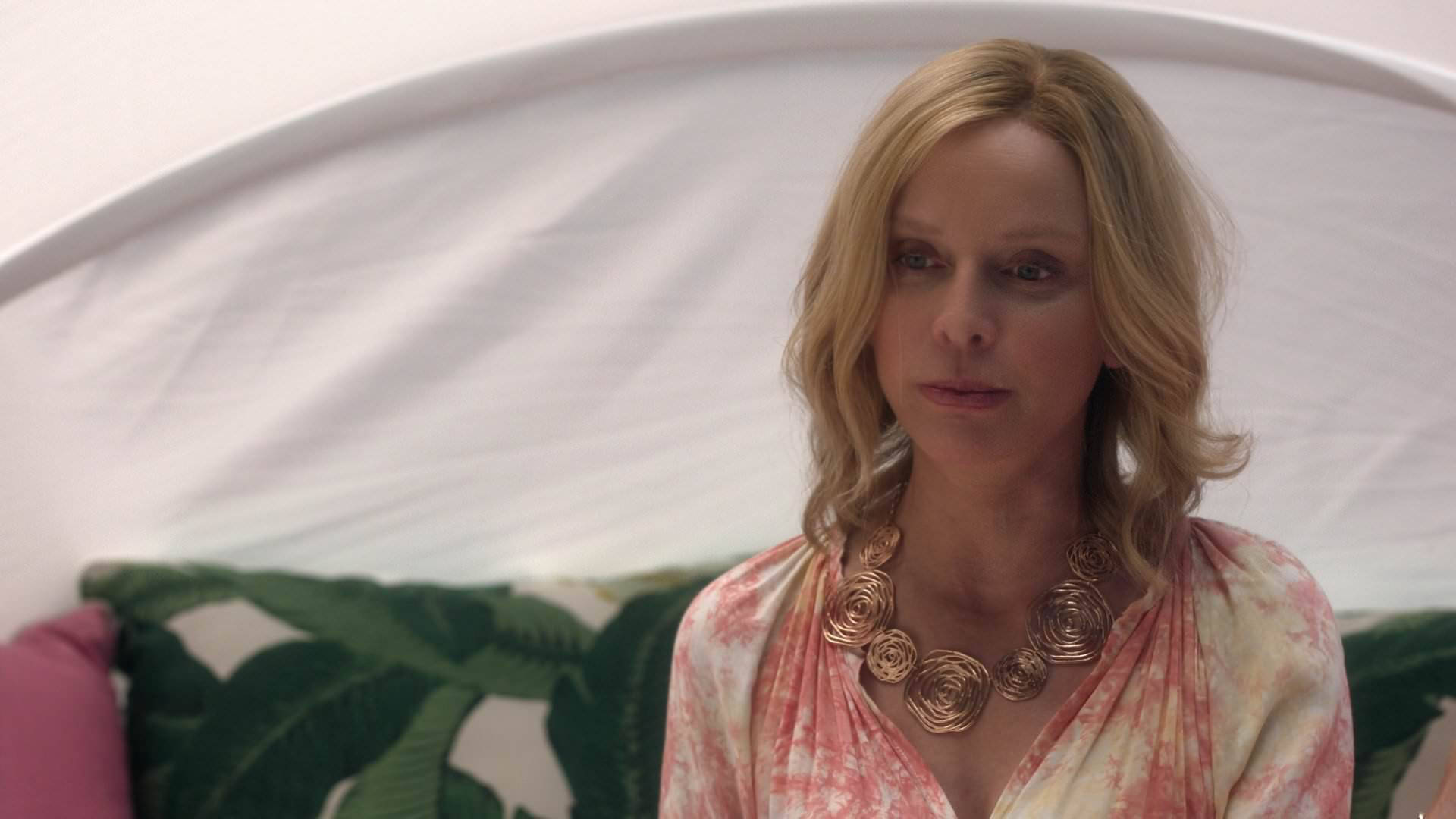Supergirl 桌布 壁紙 — 一位金髮女性穿著粉紅色紮染上衣和金色玫瑰項鍊，坐在白色天篷下的綠色葉紋沙發上。