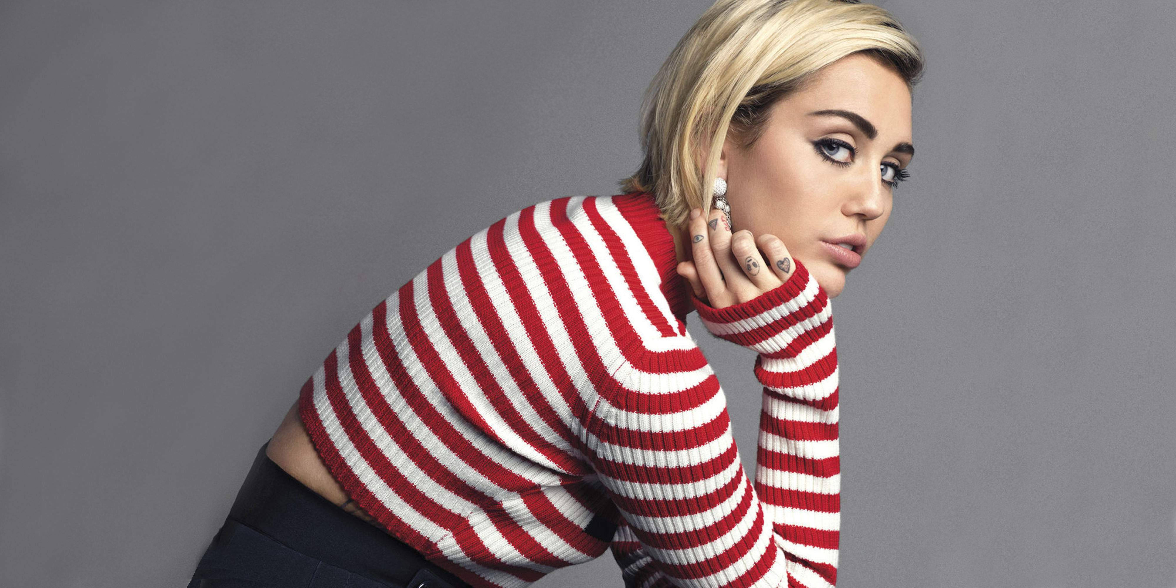 miley 桌布 壁紙 — 一位金髮女性穿著紅白條紋毛衣，手靠近臉部，在純灰色背景前擺姿勢。