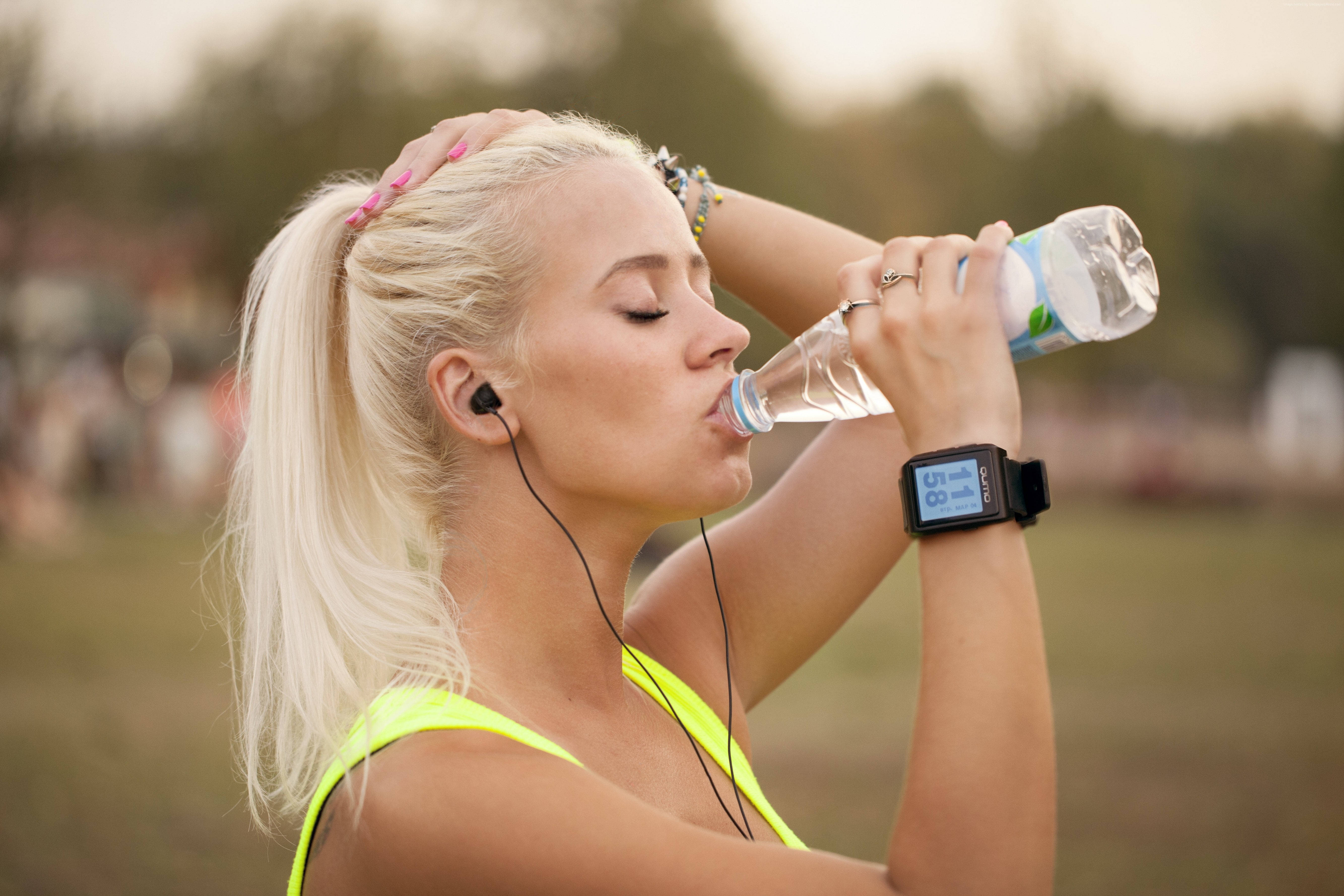Drink Water 桌布 壁紙 — 一位金髮女性穿著螢光黃運動上衣，戴著耳機和健身追蹤器，在戶外從瓶子裡喝水。