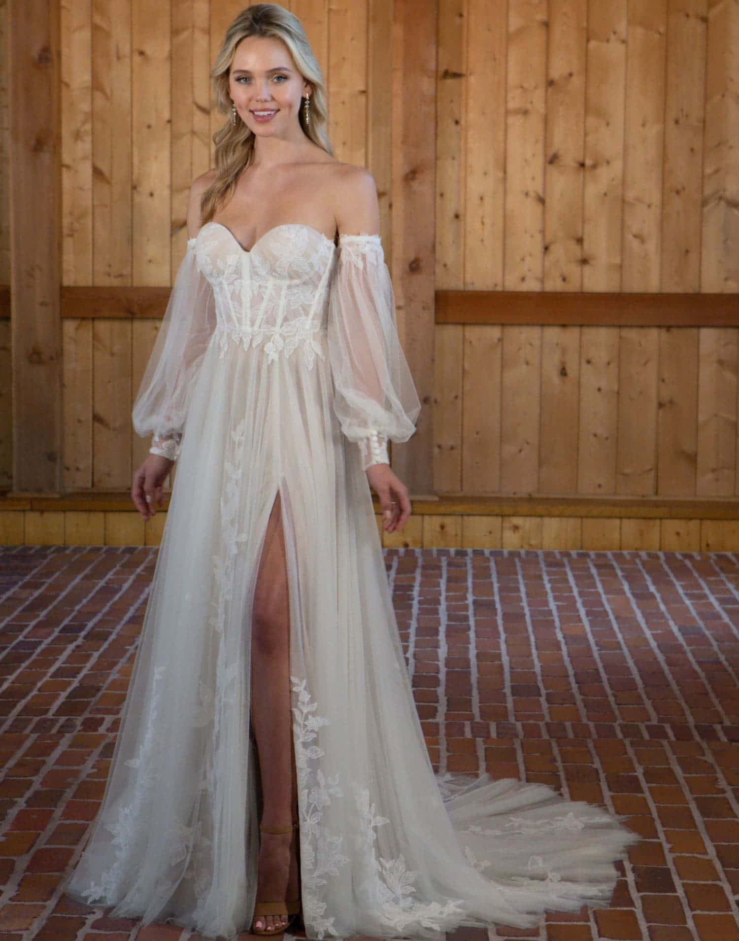 Bride's Dress 桌布 壁紙 — 一位金髮女性穿著象牙色蕾絲露肩婚紗，配以透明袖和高衩，微笑著站在木牆和磚地板前。