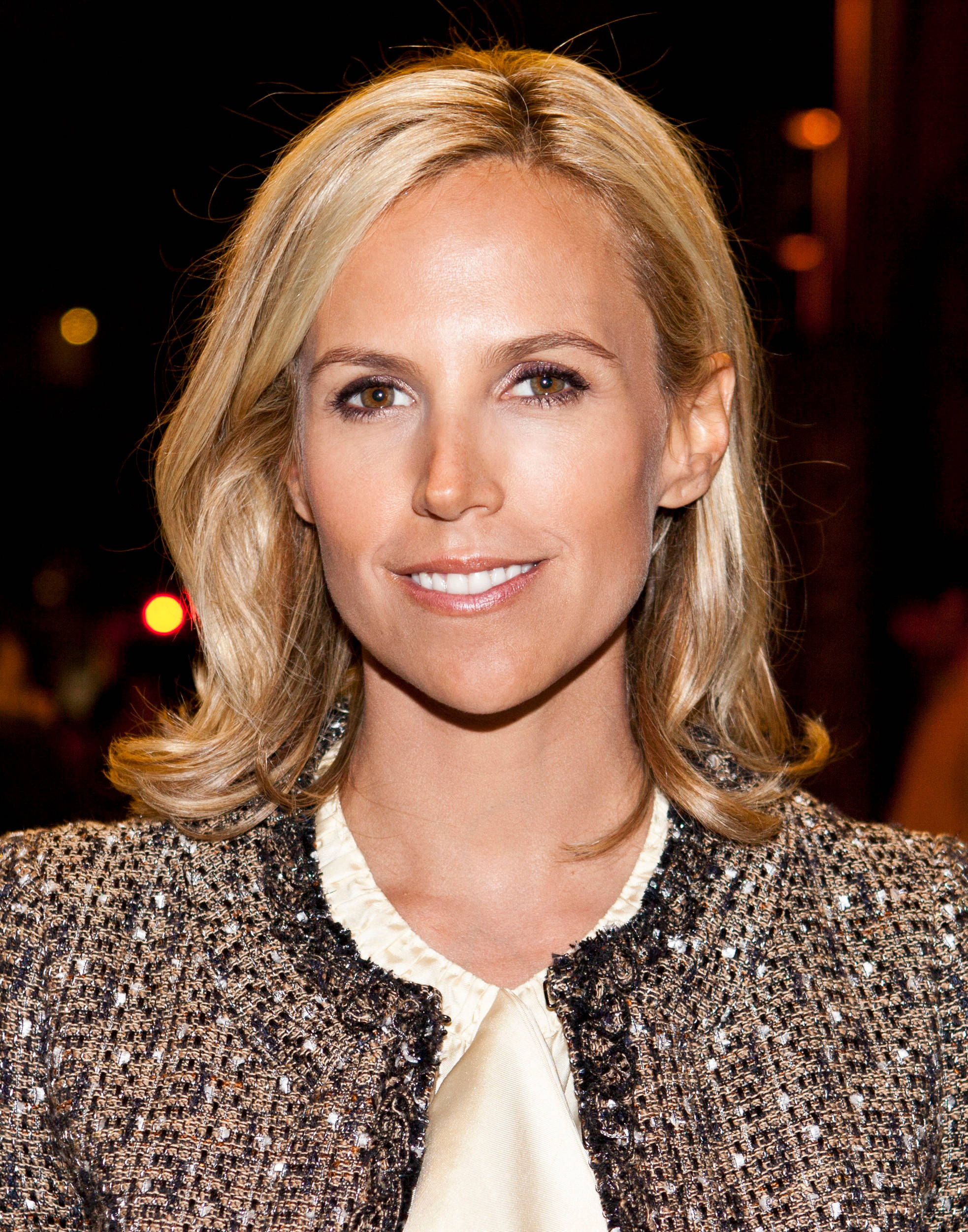 Tory Burch 桌布 壁紙 — 一位金髮女性穿著質感粗花呢外套，在模糊的夜晚背景下溫暖微笑的特寫肖像。
