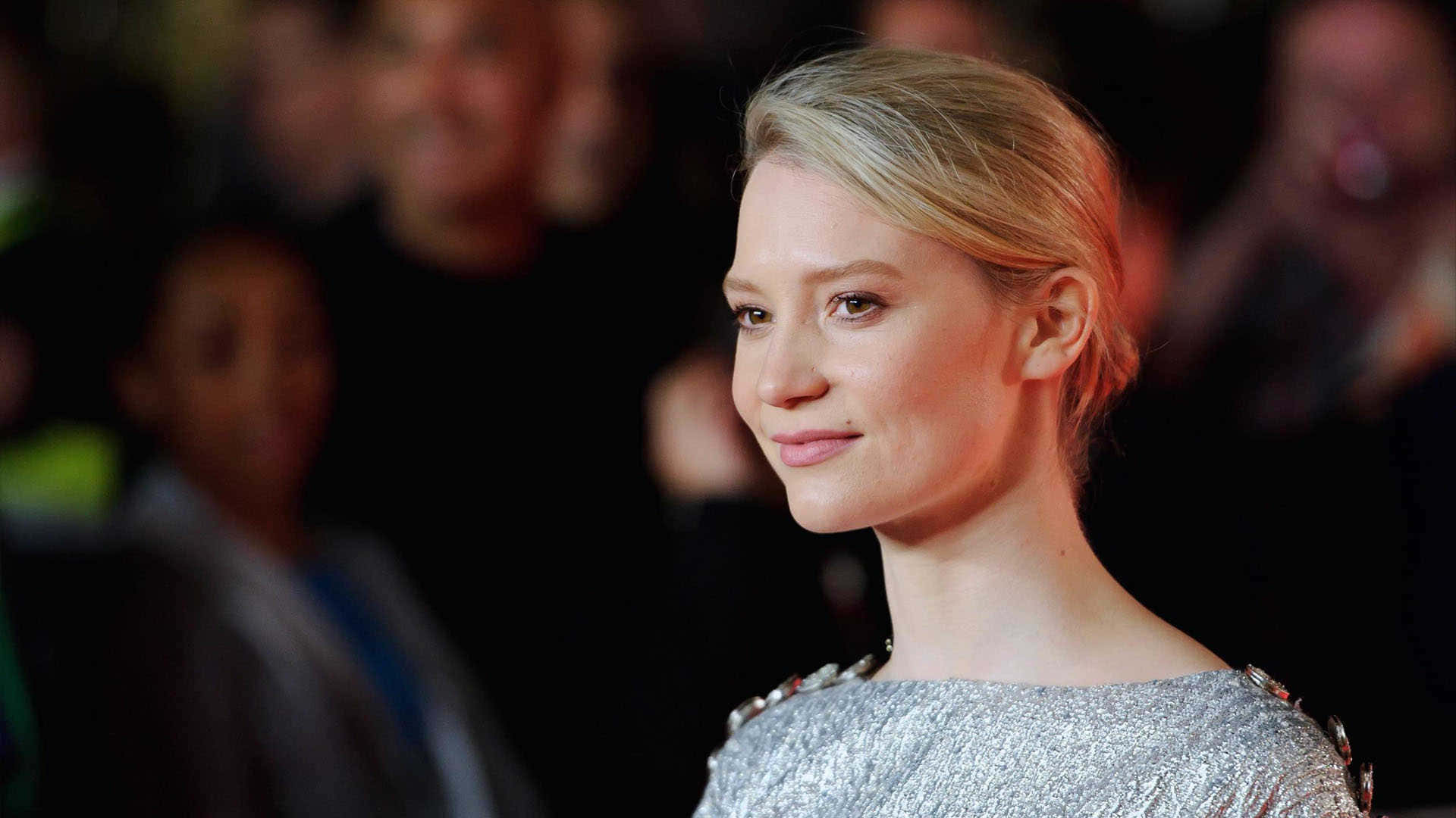 Mia Wasikowska 桌布 壁紙 — 一位金髮女性穿著銀色亮片連衣裙，在模糊人群背景的活動中溫柔微笑。