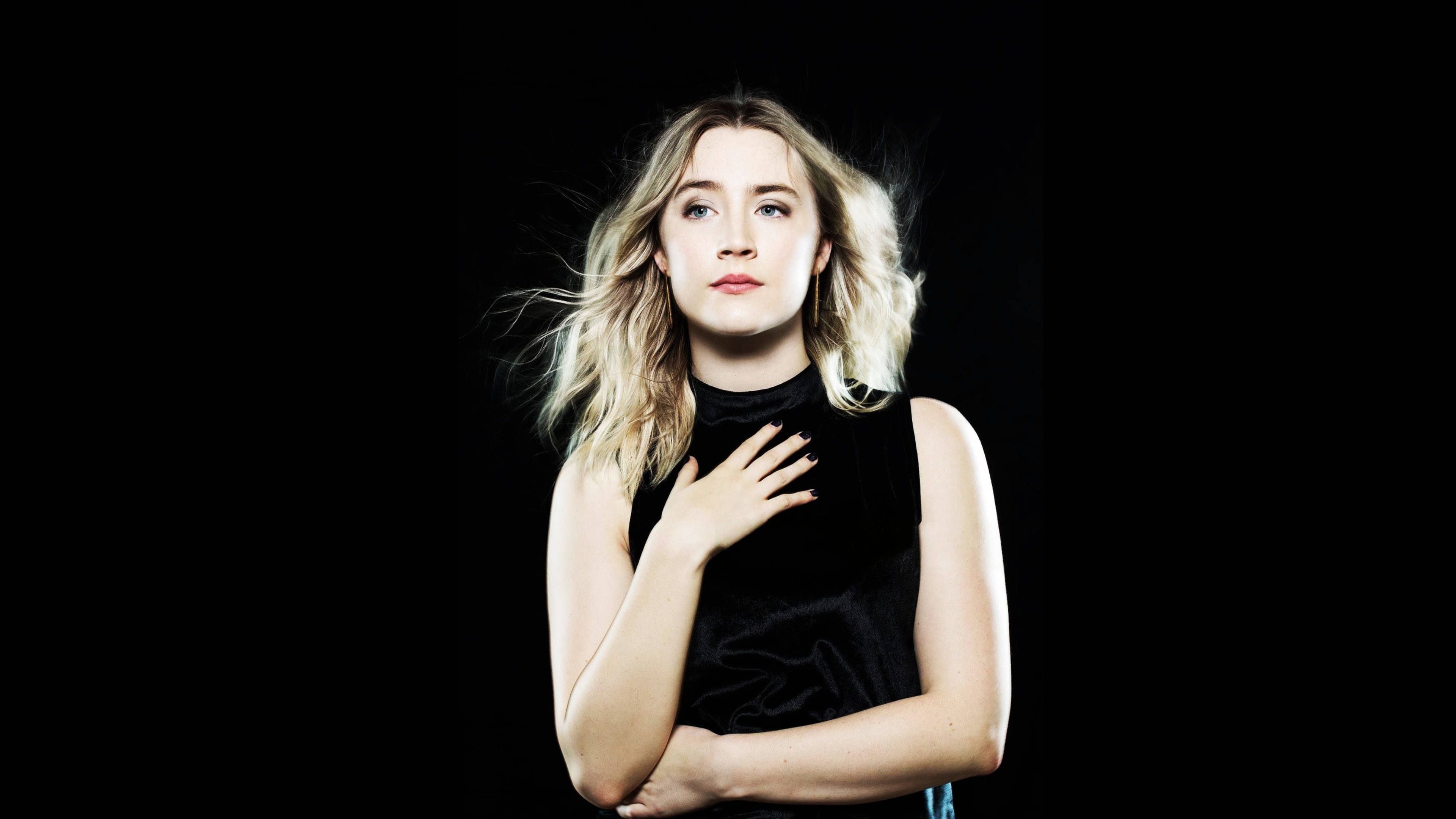 Saoirse Ronan 桌布 壁紙 — 一位金髮女性穿著黑色天鵝絨上衣，站在純黑背景前，一手放在心口，頭髮隨風飄揚。