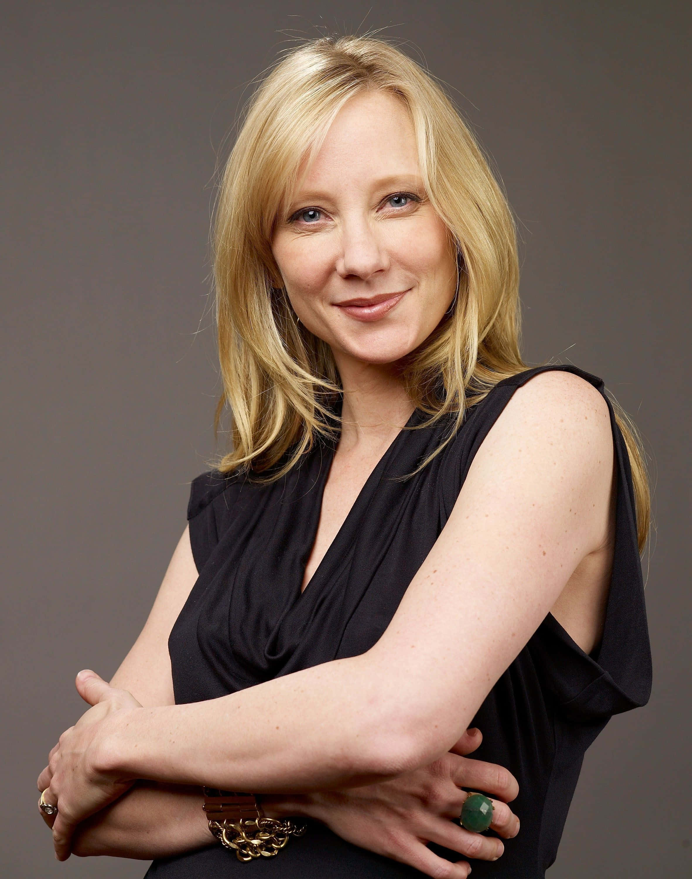 Actress HD 桌布 壁紙 — 一位金髮女性穿著黑色無袖上衣，在純灰色背景前交叉雙臂，溫暖地微笑。