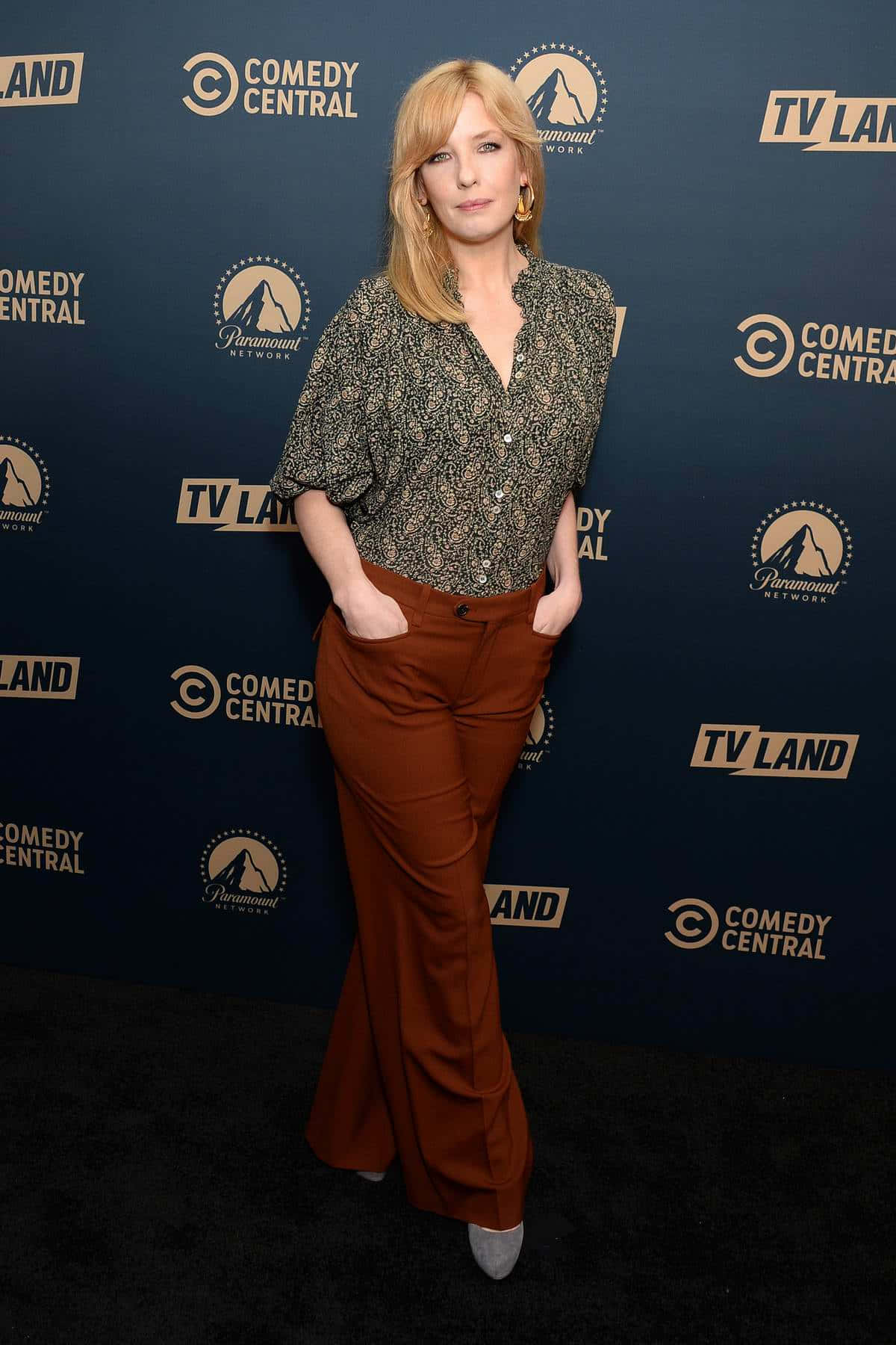 Kelly Reilly 桌布 壁紙 — 一位金髮女性自信地站在黑色地毯上，穿著圖案襯衫和鏽色喇叭褲，背景為藍色品牌背景。