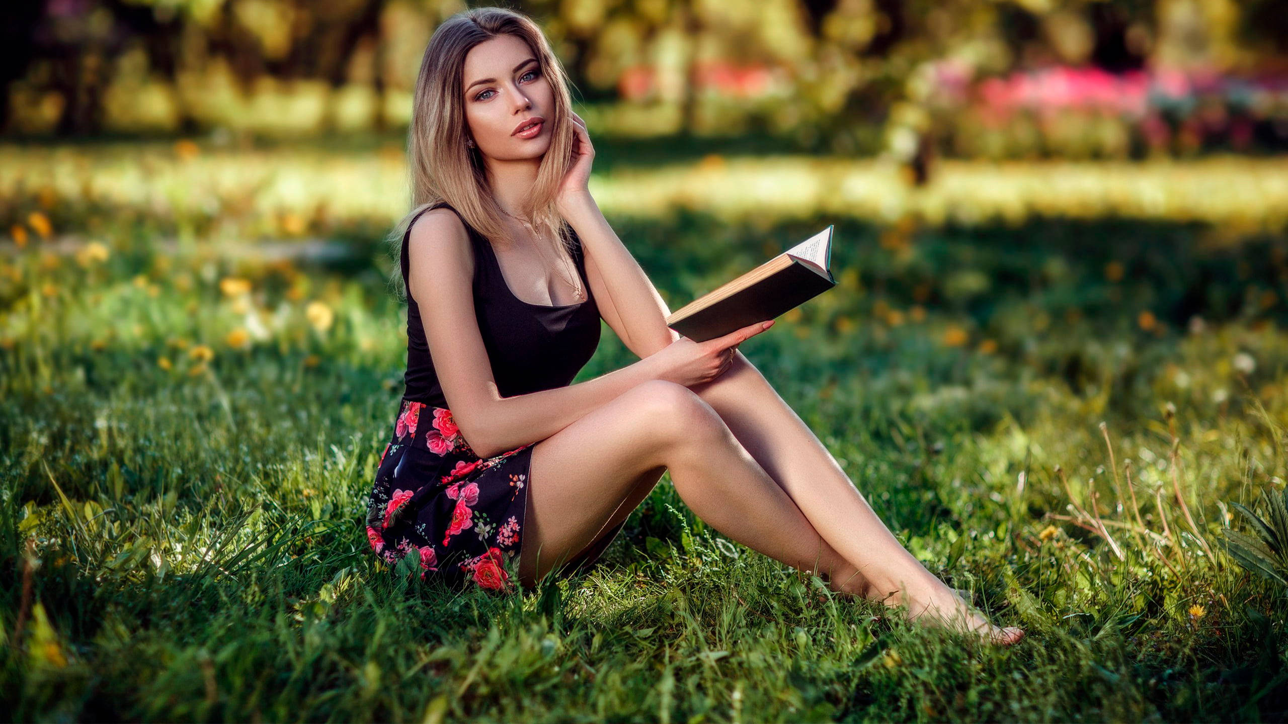 Reading 桌布 壁紙 — 一位金髮女性赤腳坐在陽光照耀的草地上，手持一本書，身穿黑色上衣和花卉圖案裙子。