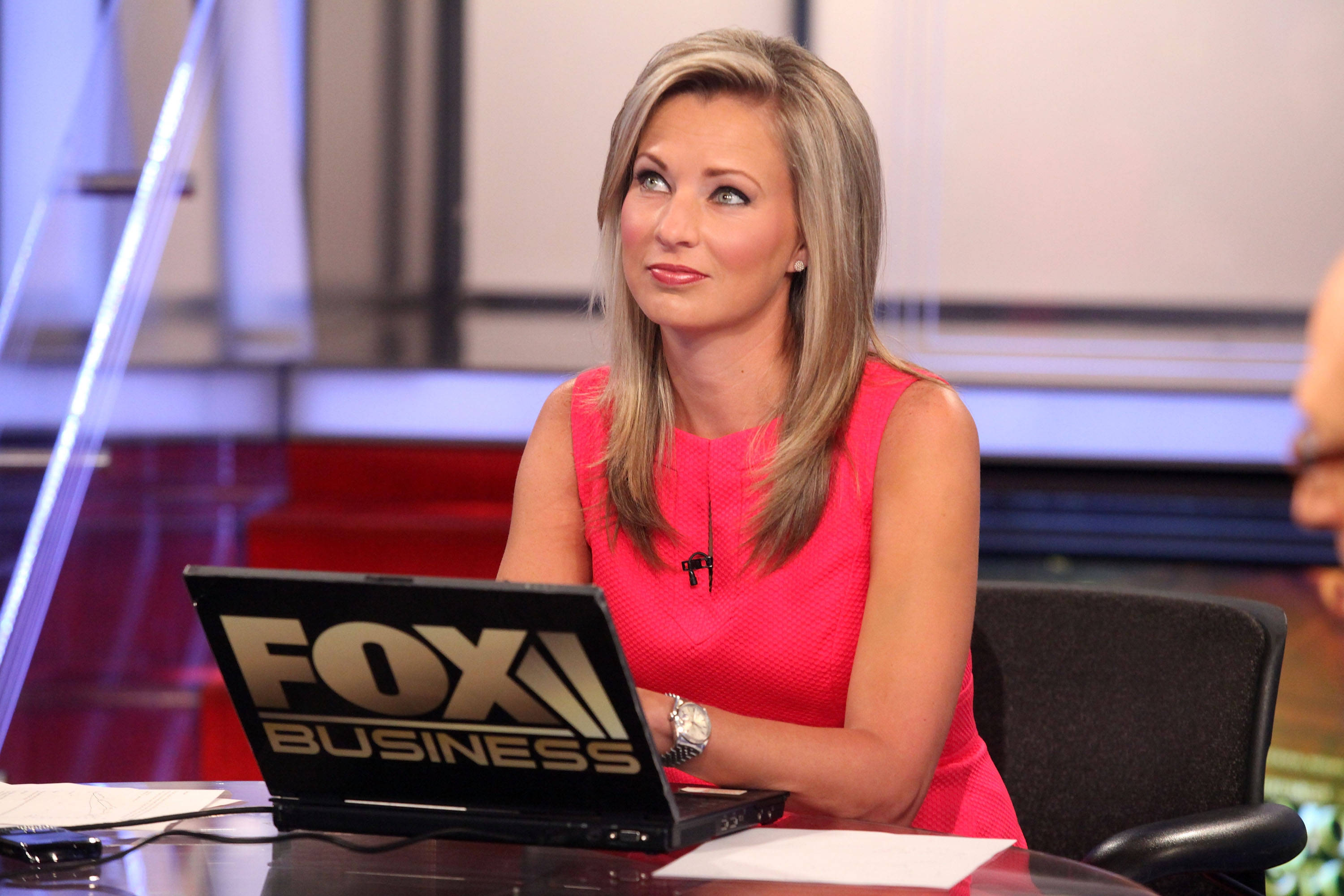News 桌布 壁紙 — 一位金髮女新聞主播穿著亮粉色連衣裙，坐在一張放有Fox Business筆記型電腦的桌子旁，視線離開鏡頭。