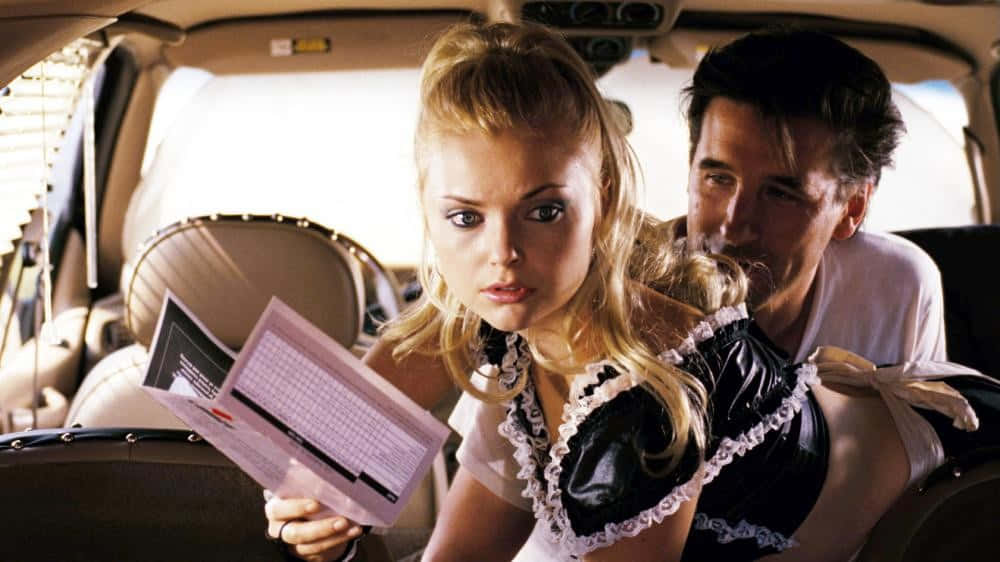 Izabella Miko 桌布 壁紙 — 一位金髮女郎穿著女傭制服，手持菜單，一名男子在車內從她身後靠過來。