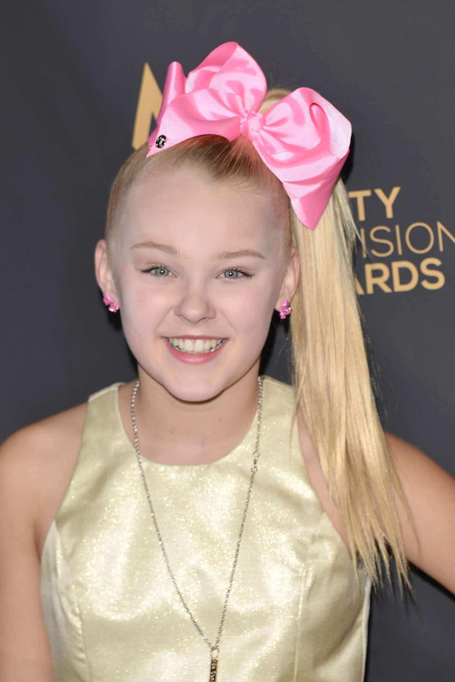 JoJo Siwa 桌布 壁紙 — 一位金髮小女孩戴著一個巨大的粉紅色蝴蝶結，穿著金色連衣裙在頒獎典禮上對著鏡頭燦爛微笑。