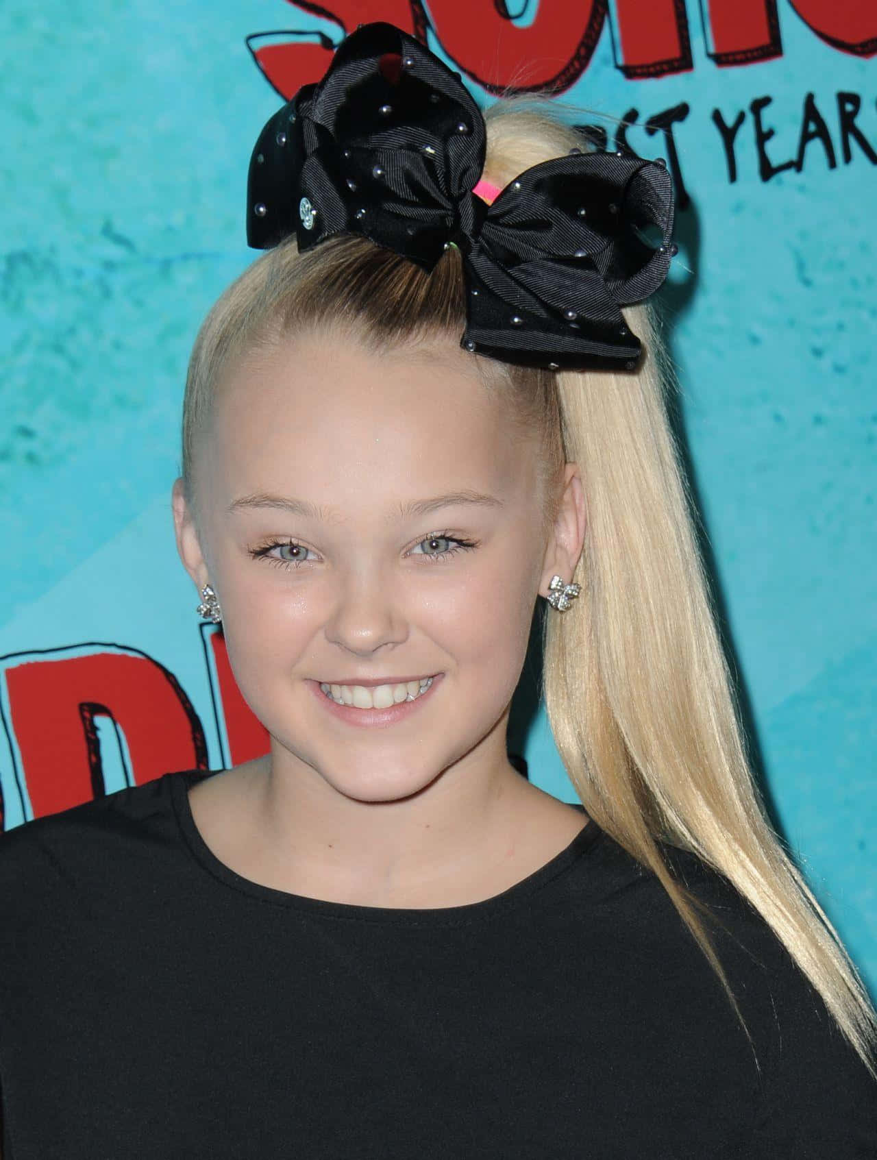 JoJo Siwa 桌布 壁紙 — 一位金髮少女戴著一個鑲有寶石的大黑色蝴蝶結，在色彩繽紛的背景前對著鏡頭燦爛微笑。
