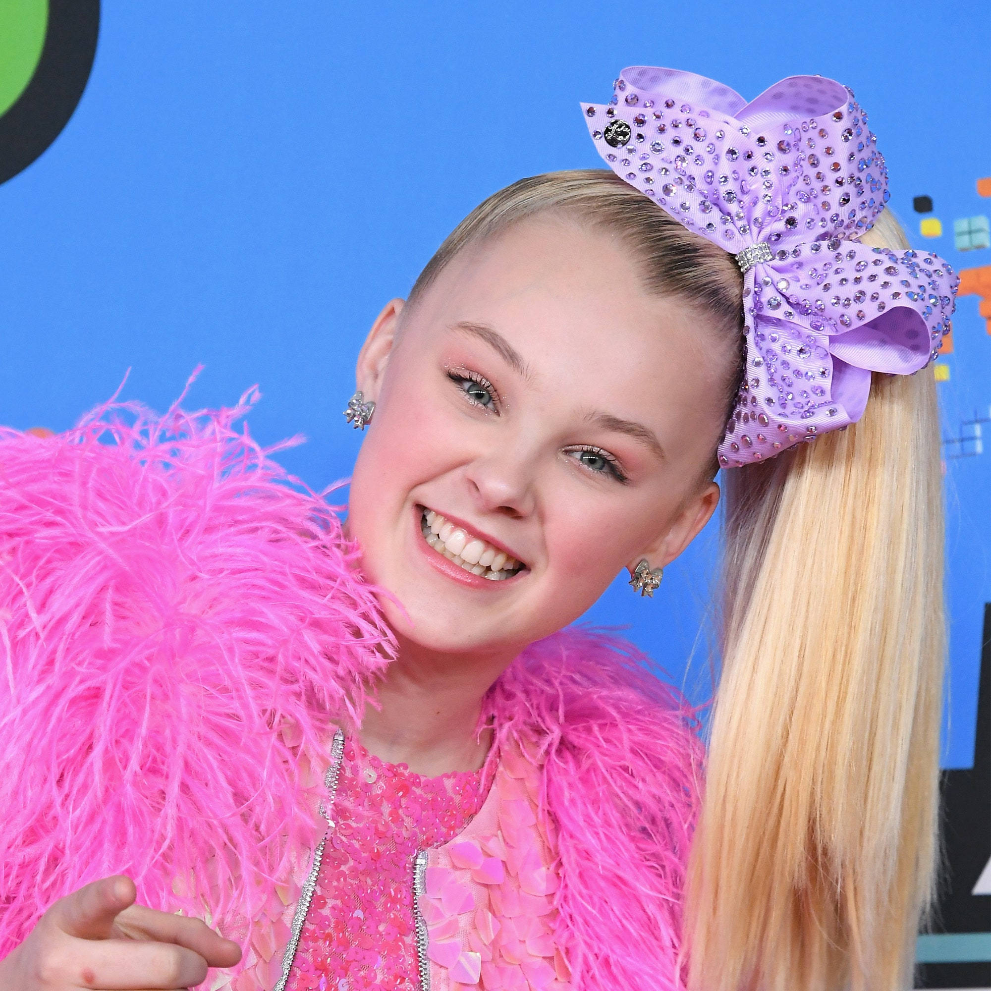 JoJo Siwa 桌布 壁紙 — 一位金髮少女戴著巨大的紫色寶石蝴蝶結，身穿鮮豔的粉紅色羽毛服裝，在藍色背景前燦爛微笑。