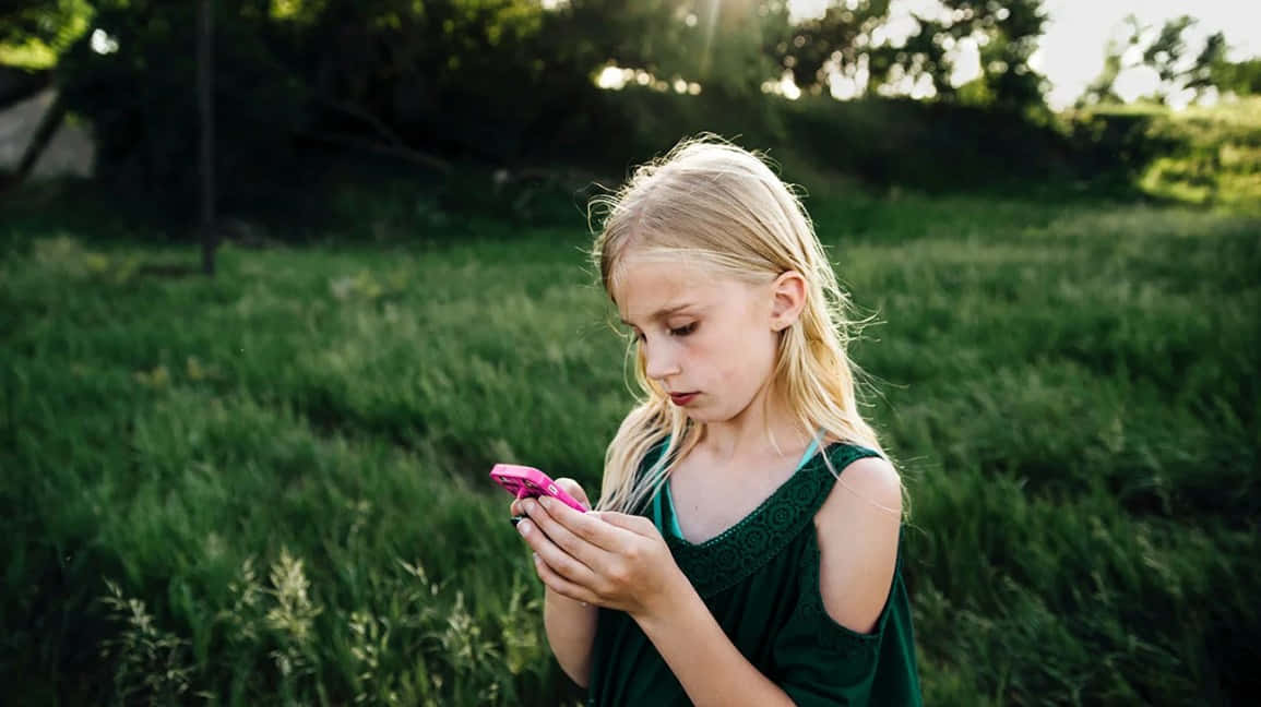 Teen Girl 桌布 壁紙 — 一位金髮少女穿著綠色連衣裙，站在陽光明媚的草地上，專注於粉紅色的手機。