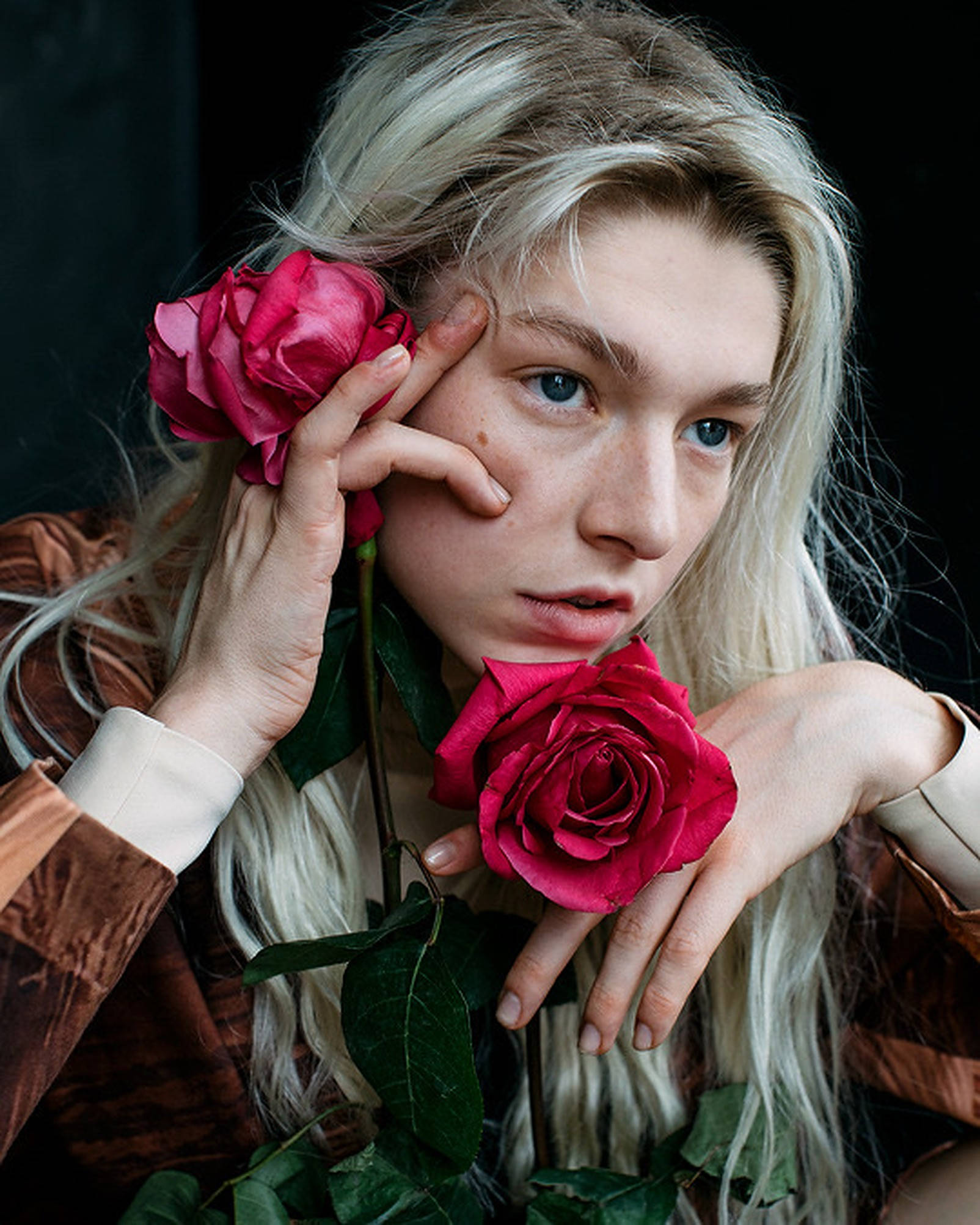 Hunter Schafer 桌布 壁紙 — 一位金髮年輕人在黑暗背景下手持鮮豔的紅玫瑰，營造出充滿氛圍的肖像。