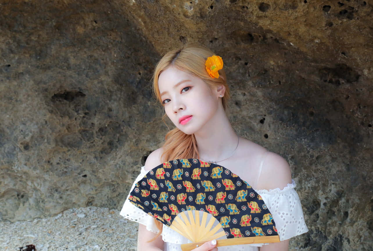 twice 桌布 壁紙 — 一位金髮年輕女子，頭髮上插著一朵橙色花，手持裝飾扇，靠著紋理岩壁。