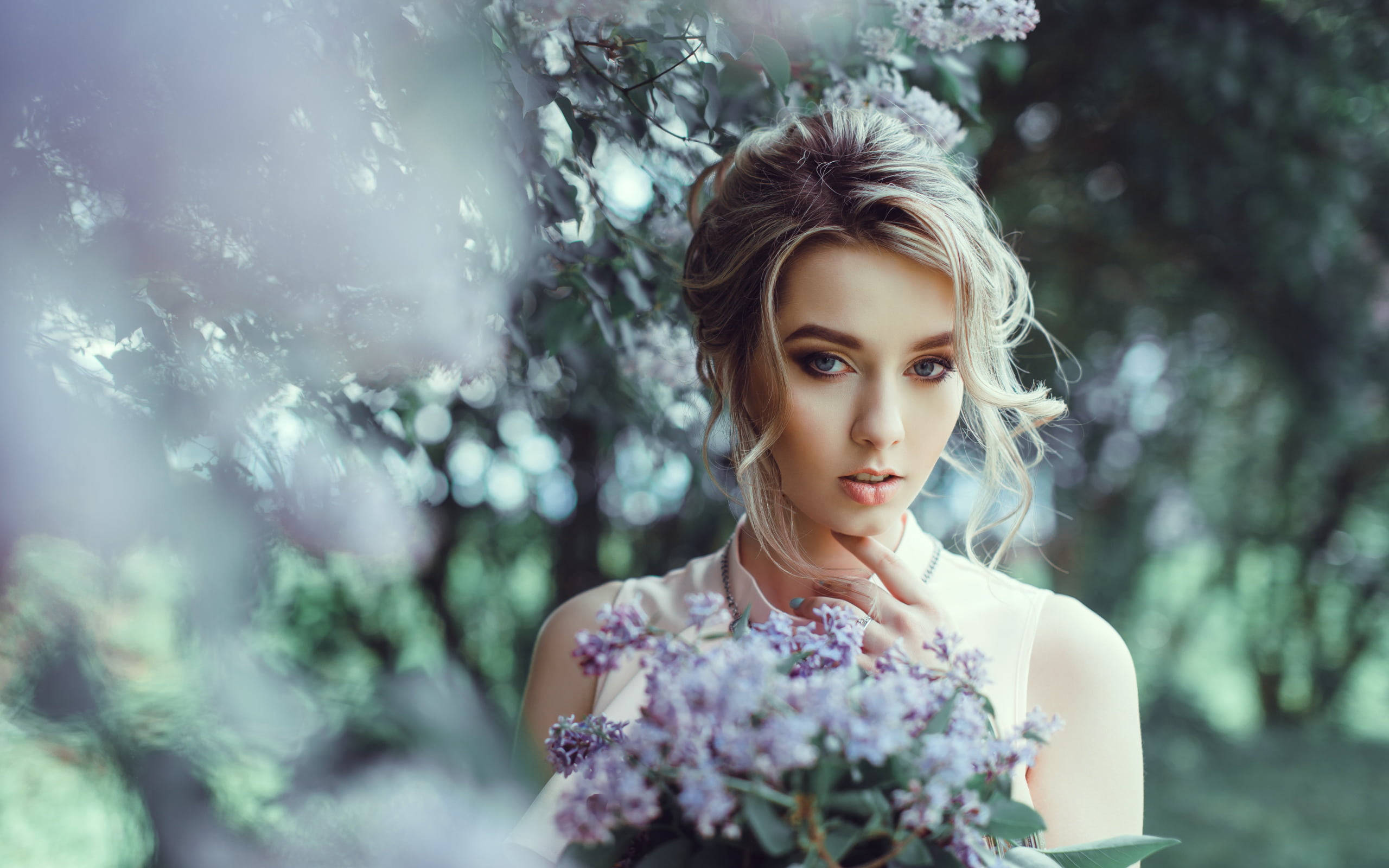 Beautiful Woman 桌布 壁紙 — 一位金髮年輕女子在柔焦花園中，於一棵開花的樹下拿著一束紫色丁香花。