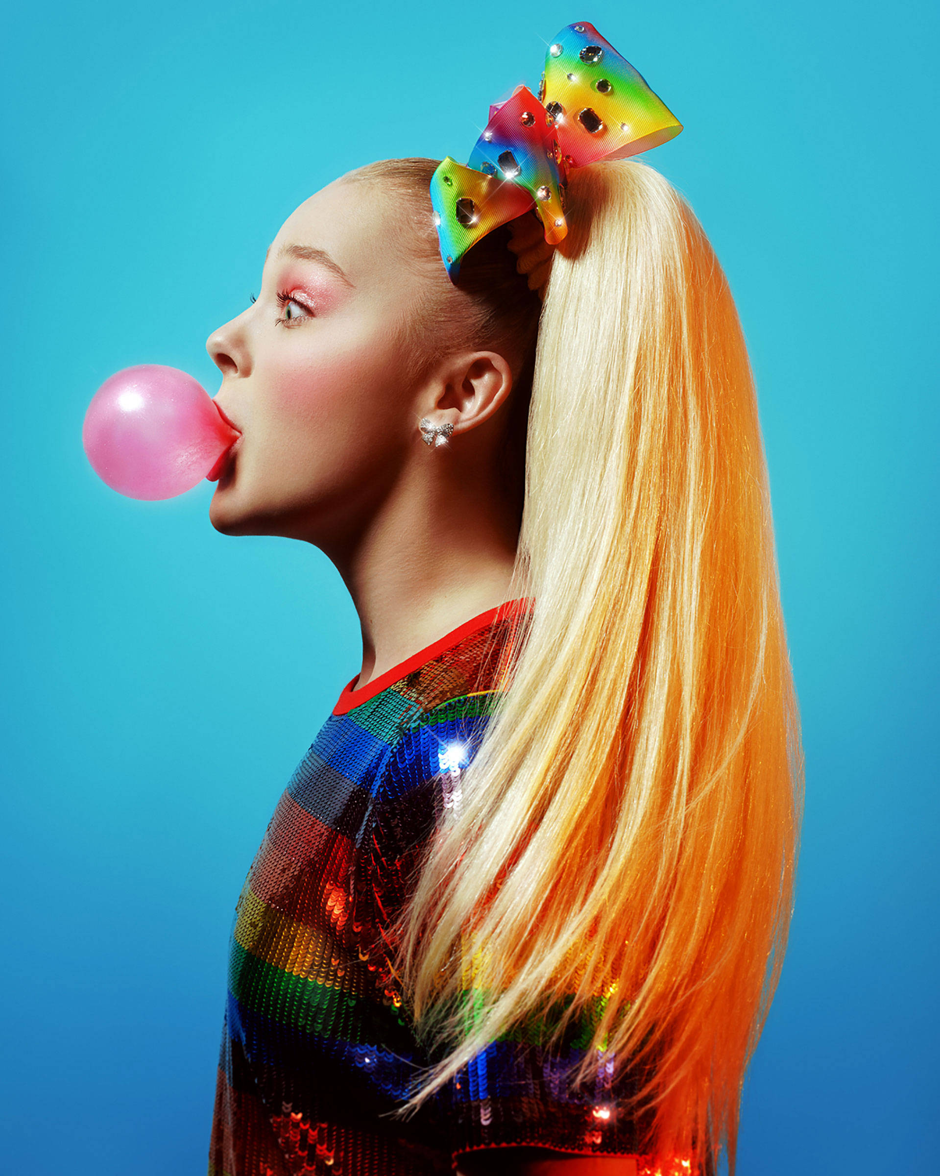 JoJo Siwa 桌布 壁紙 — 一位金髮年輕女子戴著彩虹蝴蝶結，在鮮豔的藍色背景下吹出粉紅色泡泡糖泡泡。