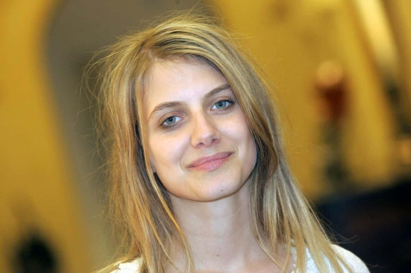 Mélanie Laurent 桌布 壁紙 — 一位金髮年輕女子的特寫肖像，帶著溫柔的微笑，背景是溫暖的金色模糊背景。