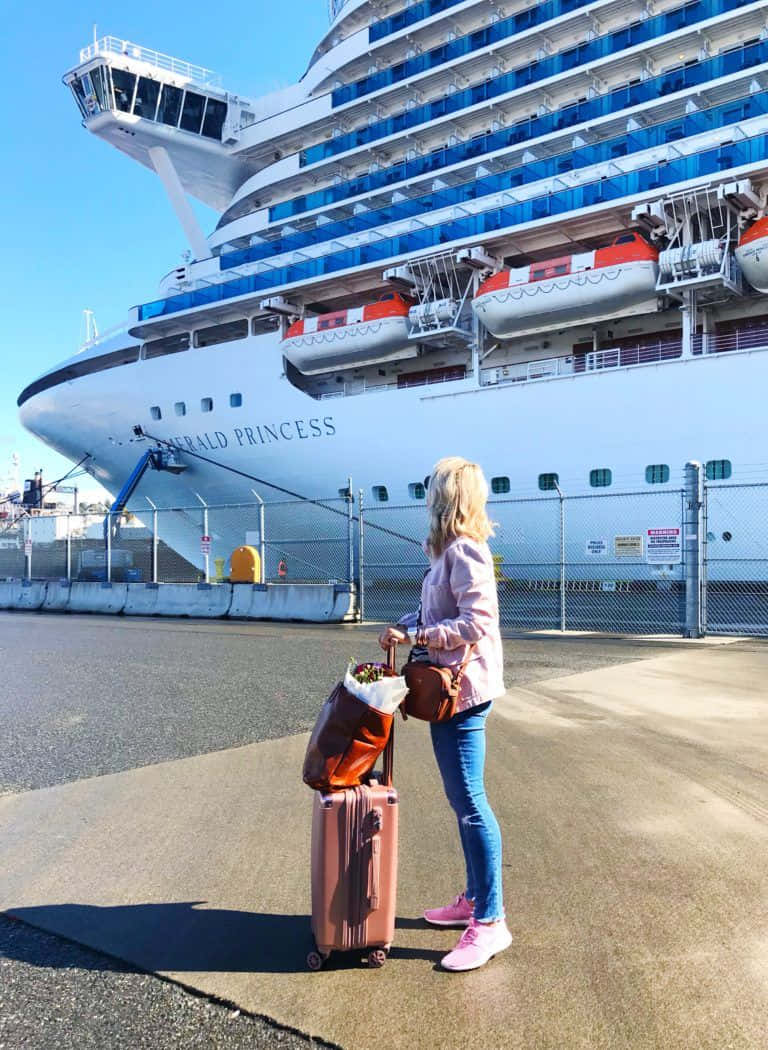 Cruise 桌布 壁紙 — 一位金髮年輕女子站在一個粉紅色行李箱旁邊，面前是停泊在陽光明媚港口的巨大白色和藍色郵輪翡翠公主號。