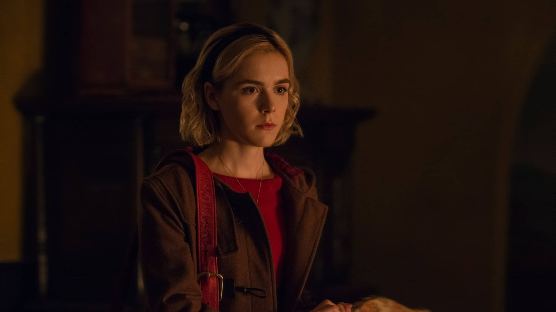 Kiernan Shipka 桌布 壁紙 — 一位金髮年輕女性，背著紅色肩帶包，站在光線昏暗、溫暖的房間裡，目光離開相機，表情嚴肅。