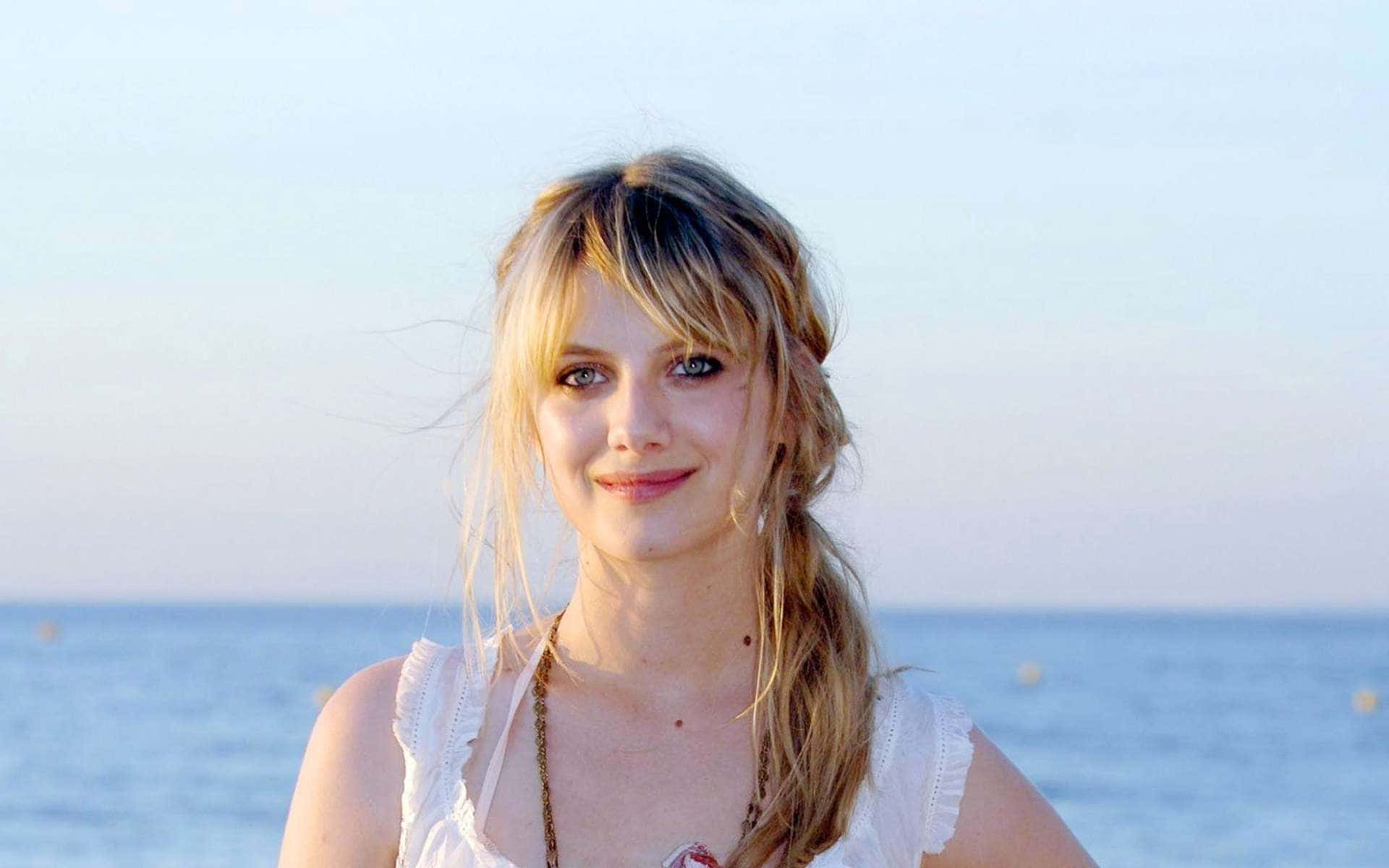 Mélanie Laurent 桌布 壁紙 — 一位金髮年輕女性在柔和日光下，以平靜的海洋為背景，對著鏡頭溫柔微笑。