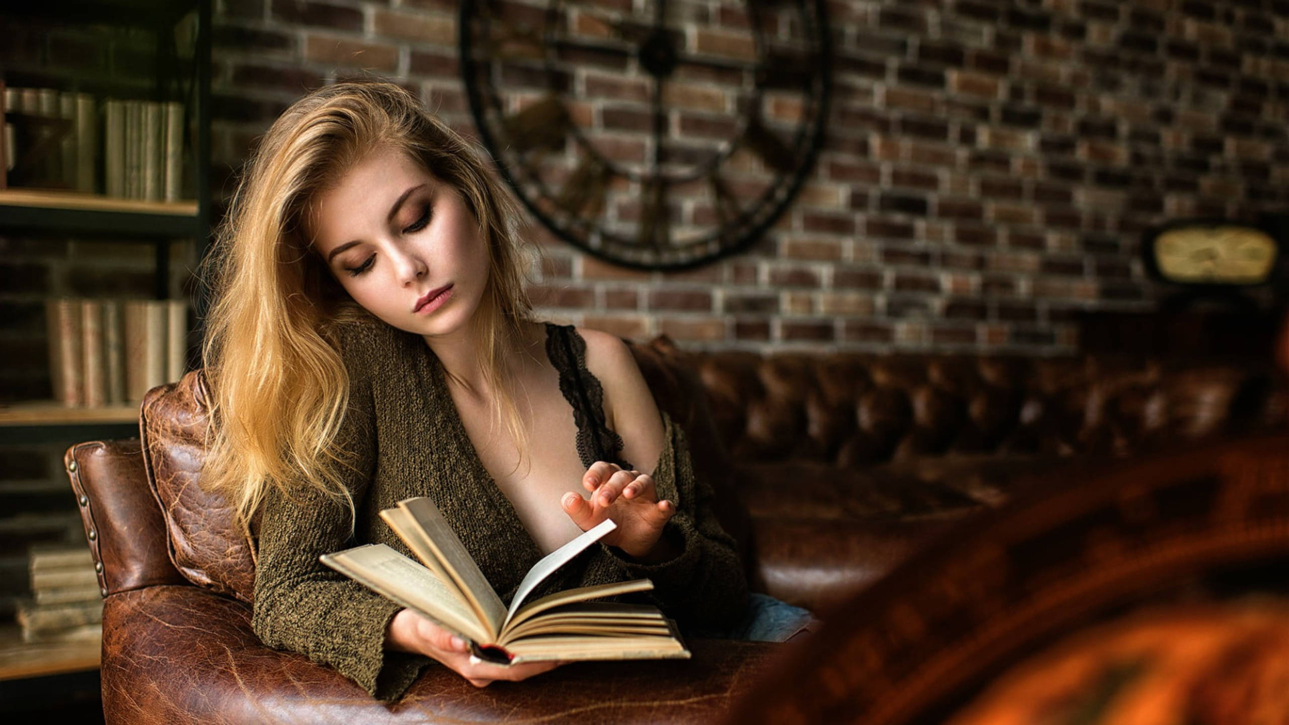 Reading 桌布 壁紙 — 一位金髮年輕女性坐在皮革扶手椅上，在一個有磚牆、書架和大時鐘的舒適房間裡看書。