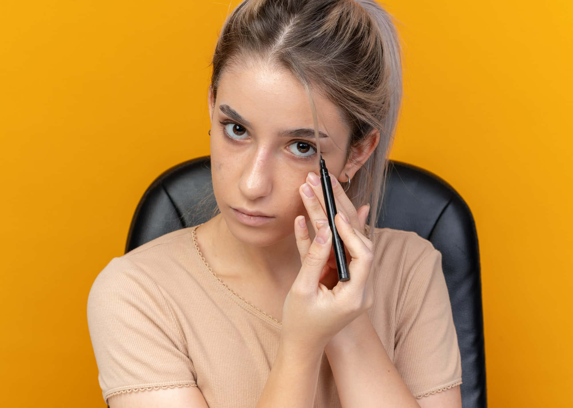 美女 桌布 壁紙 — 一位金髮年輕女性坐在黑色椅子上，背景是充滿活力的橙色牆壁，她手持黑色眼線筆靠近眼睛，表情專注。