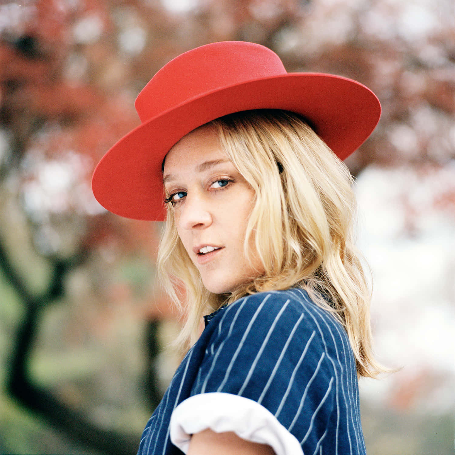 Chloë Sevigny 桌布 壁紙 — 一位金髮年輕女性戴著鮮紅色帽子，穿著藍色細條紋襯衫，背景是柔和模糊的秋日樹木。