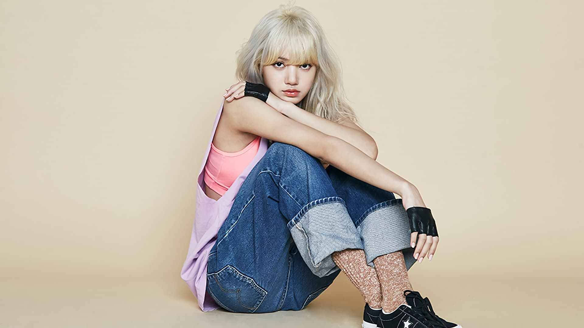 Lalisa 桌布 壁紙 — 一位金髮年輕女性盤腿坐在米色地板上，穿著牛仔褲、粉紅色上衣和黑色運動鞋。