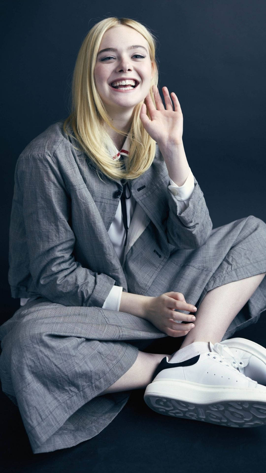 Elle 桌布 壁紙 — 一位金髮年輕女性盤腿而坐，穿著灰色西裝和白色運動鞋，微笑著向鏡頭揮手。