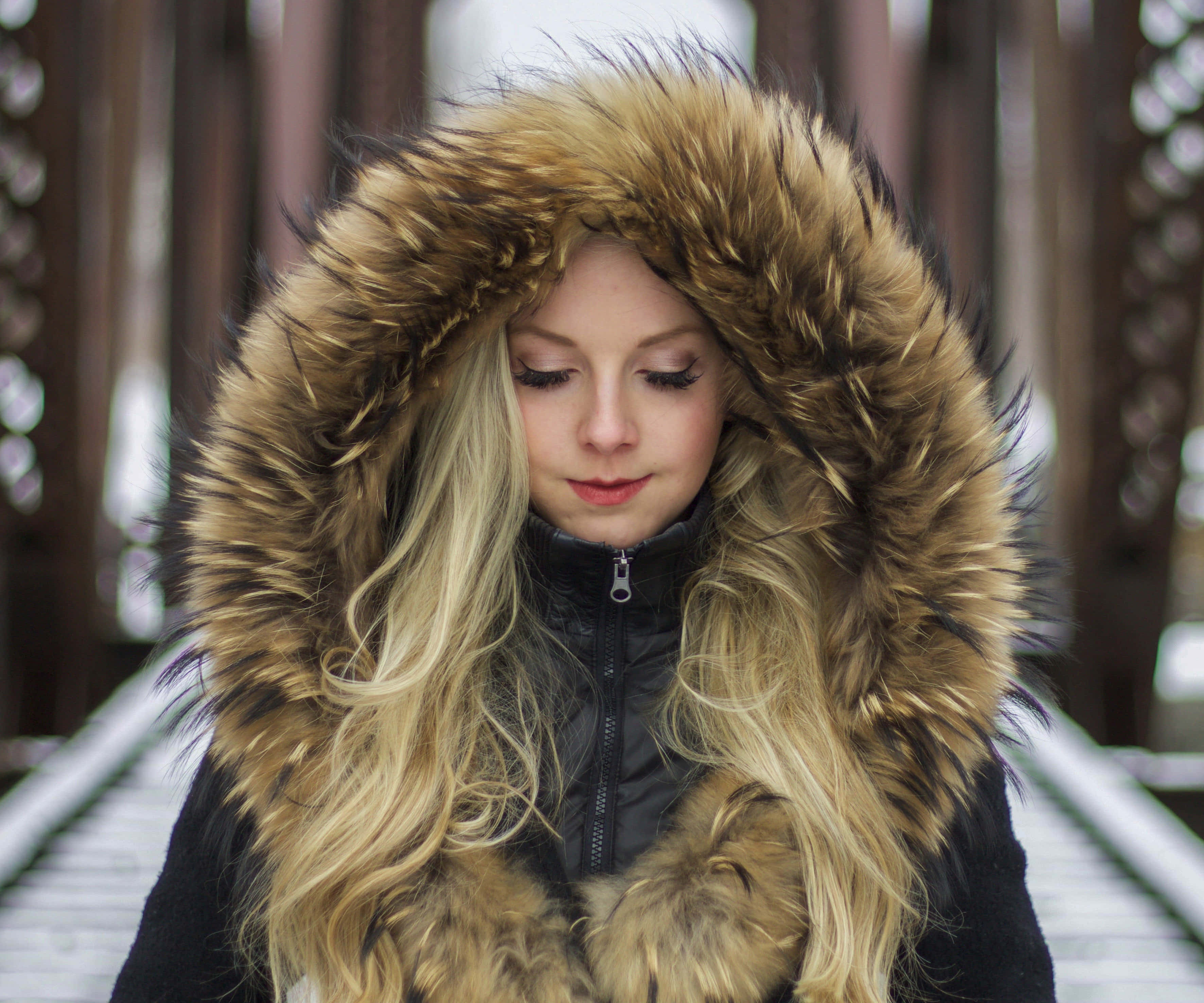 Animal Fur 桌布 壁紙 — 一位金髮年輕女性穿著黑色冬季外套，配有大型棕色毛絨內襯連帽，站在木橋上。