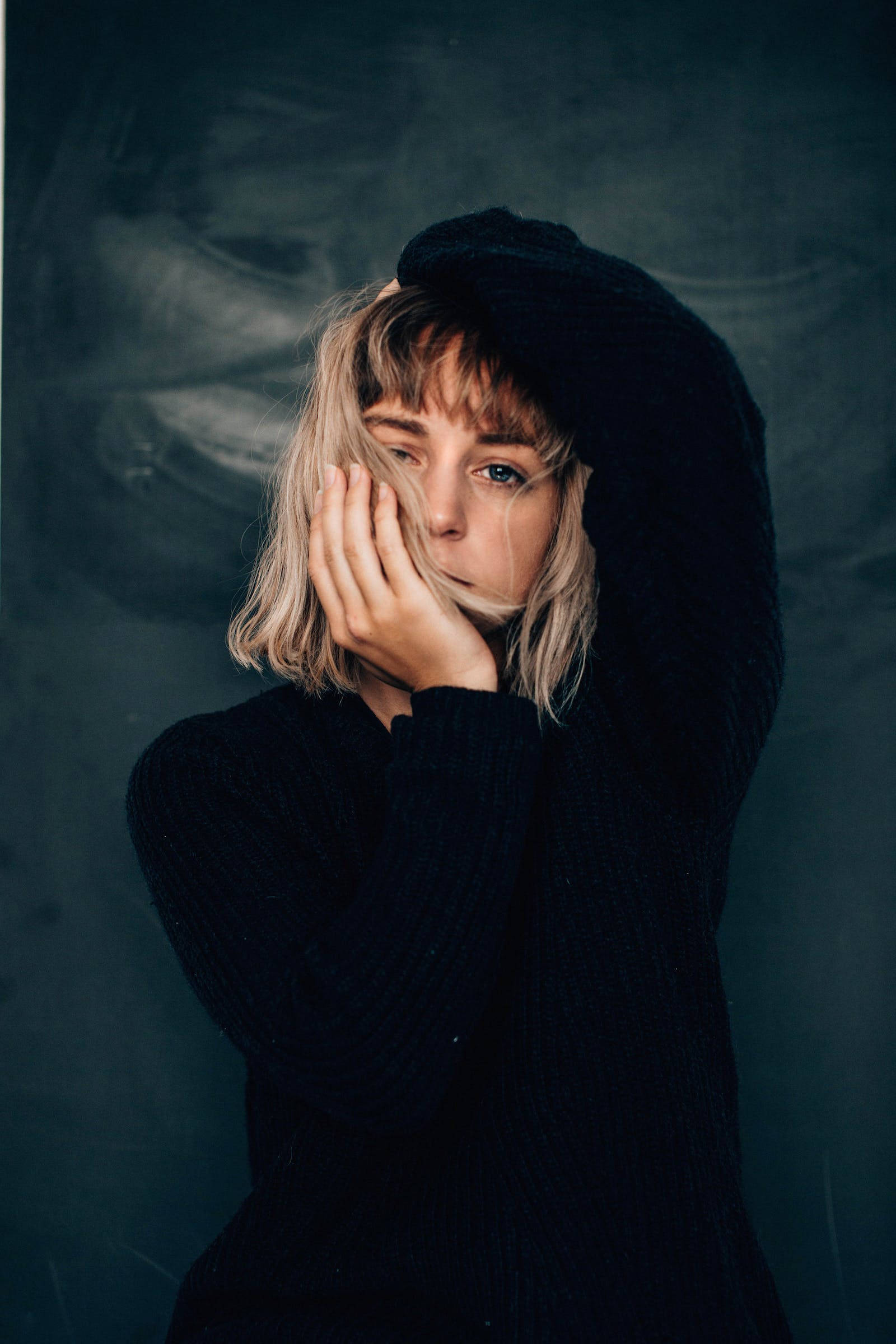 Cool Sad 桌布 壁紙 — 一位金髮年輕女性穿著黑色連帽衫，在深色紋理背景前輕輕用手遮住臉。