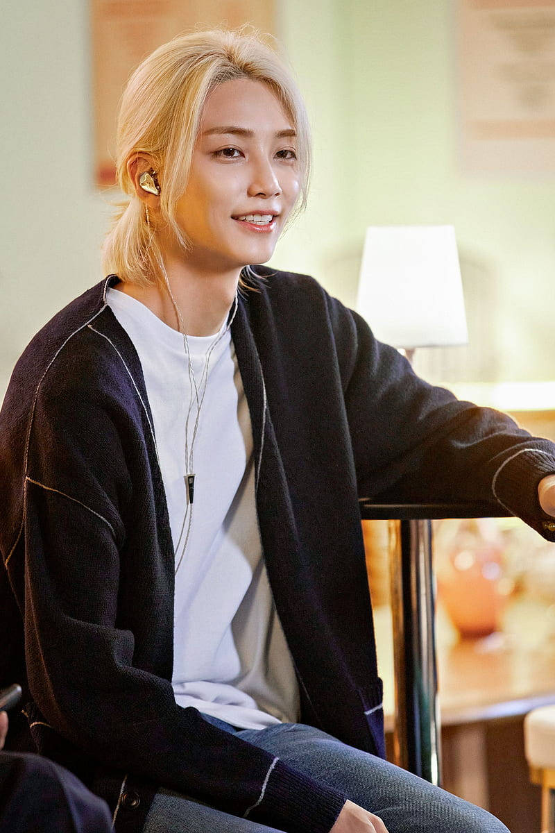 yoon jeonghan 桌布 壁紙 — 一位金髮年輕男子在室內戴著耳塞和黑色開襟衫微笑。