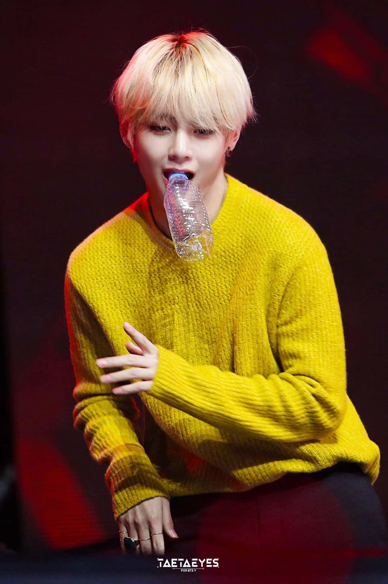 Taehyung Cute 桌布 壁紙 — 一位金髮年輕男子在舞台燈光下穿著亮黃色針織毛衣，從透明瓶子裡喝水。