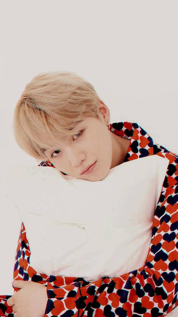 Suga Bts 桌布 壁紙 — 一位金髮年輕男子將頭靠在白色枕頭上，身穿紅藍心形圖案睡袍，背景為純白色。