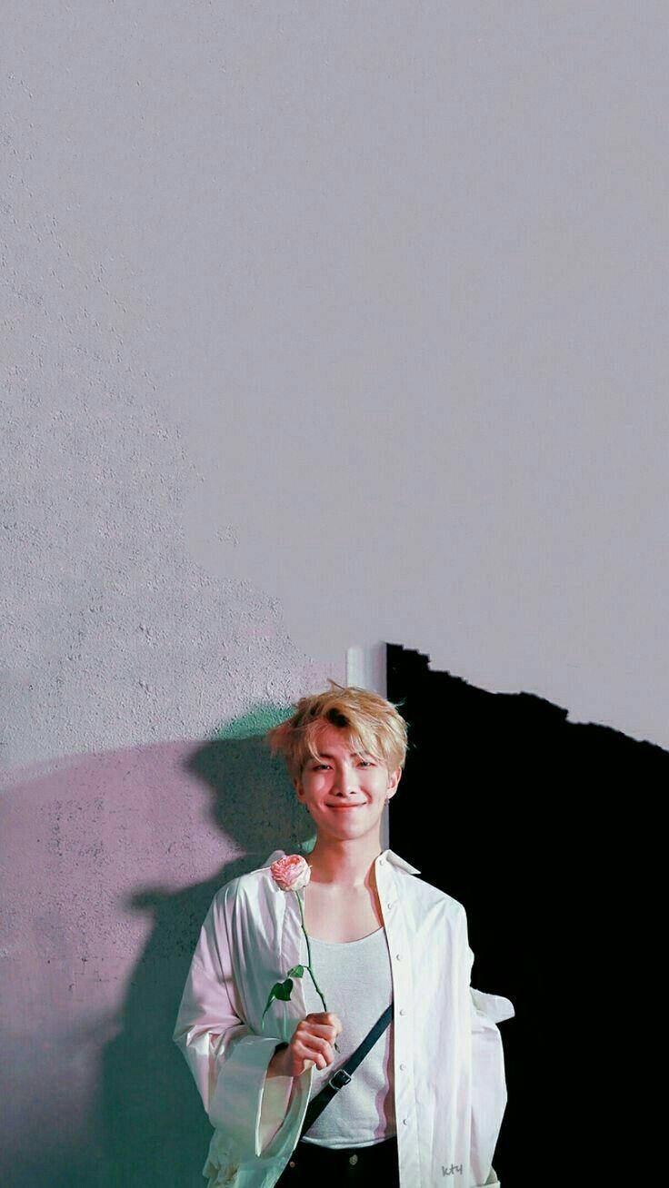 Namjoon 桌布 壁紙 — 一位金髮年輕男子微笑著，手持粉紅玫瑰，背景是純白牆壁，帶有戲劇性的陰影。