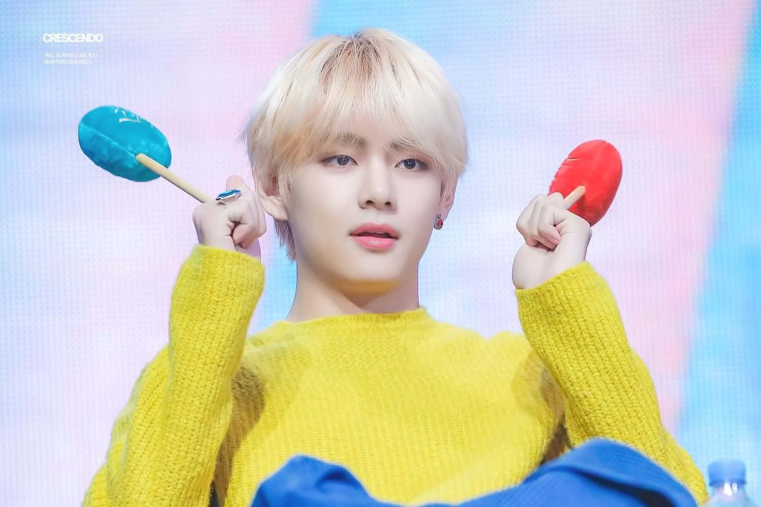 Taehyung Cute 桌布 壁紙 — 一位金髮年輕男子穿著亮黃色毛衣，在柔和漸變背景前手持彩色棒棒糖。