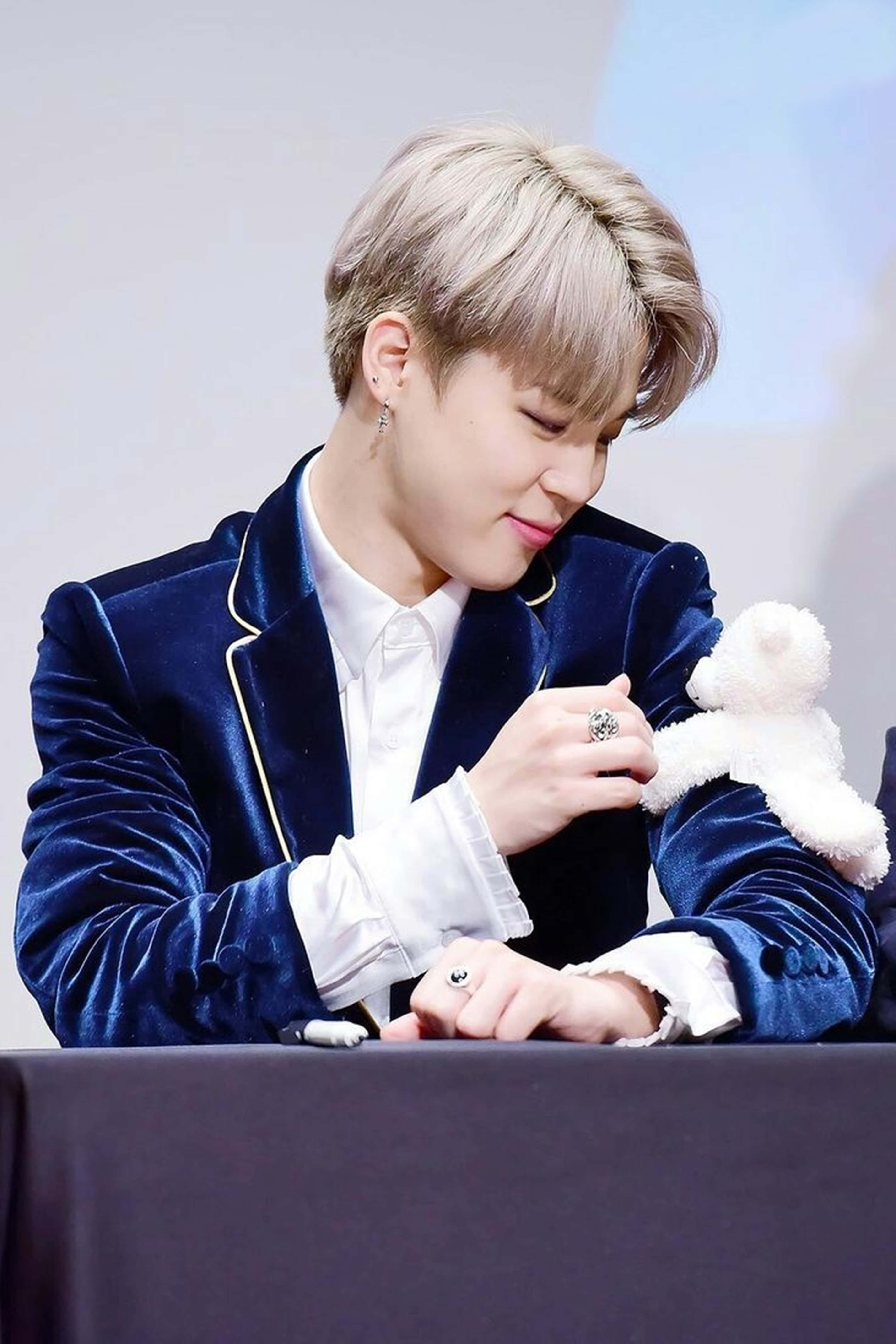 BTS 桌布 壁紙 — 一位金髮年輕男子穿著藍色天鵝絨西裝外套，在簽名活動中溫柔地為一隻白色泰迪熊簽名。