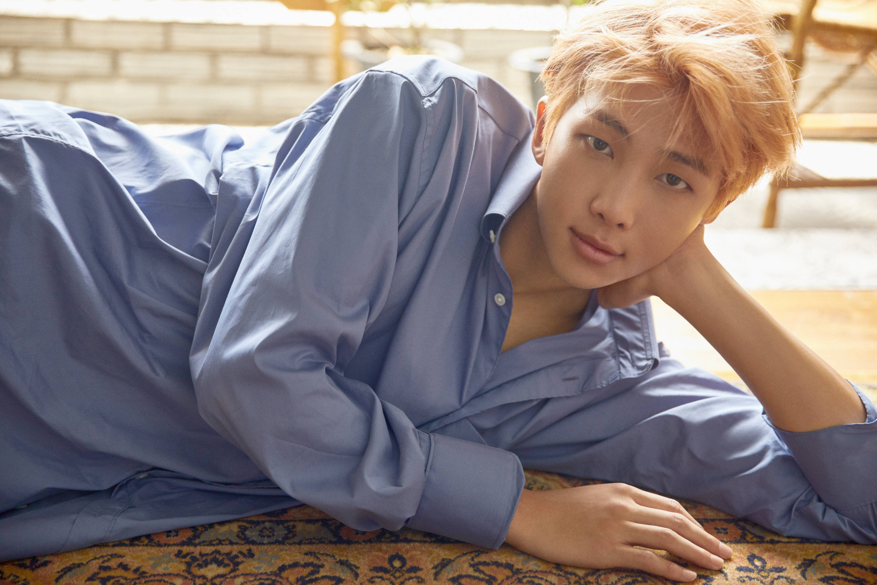 BTS 桌布 壁紙 — 一位金髮年輕男子穿著藍色襯衫，放鬆地躺在圖案地毯上，溫柔地凝視鏡頭。