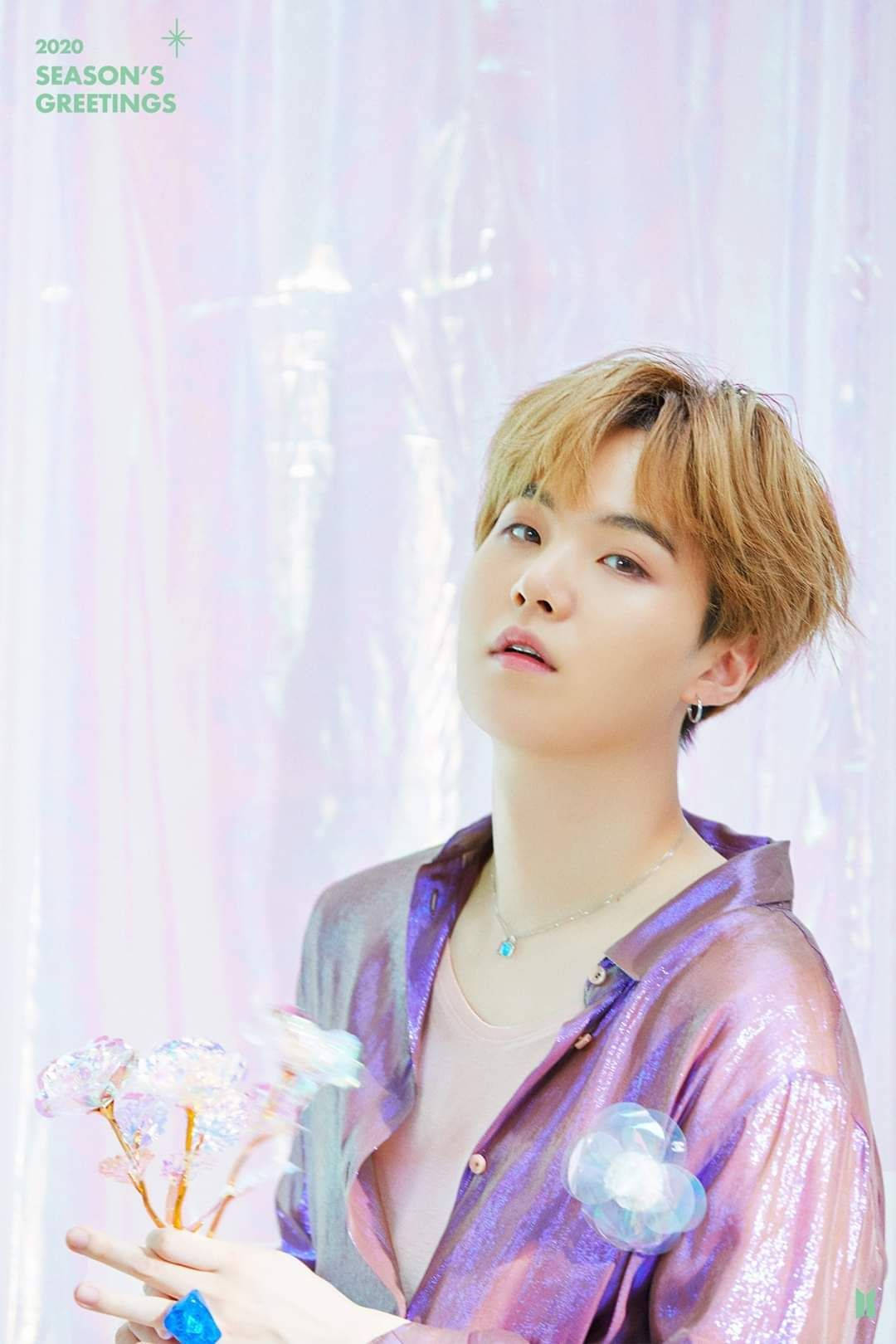 BTS 桌布 壁紙 — 一位金髮年輕男子穿著閃亮紫色襯衫，在柔和的粉紅色和白色背景前握著發光的花朵。