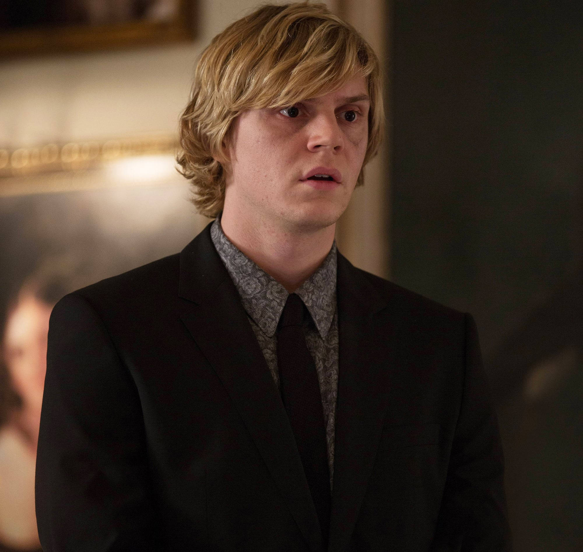 Tate Langdon 桌布 壁紙 — 一位金髮年輕男子穿著黑色西裝和領帶，在室內露出驚訝的表情。
