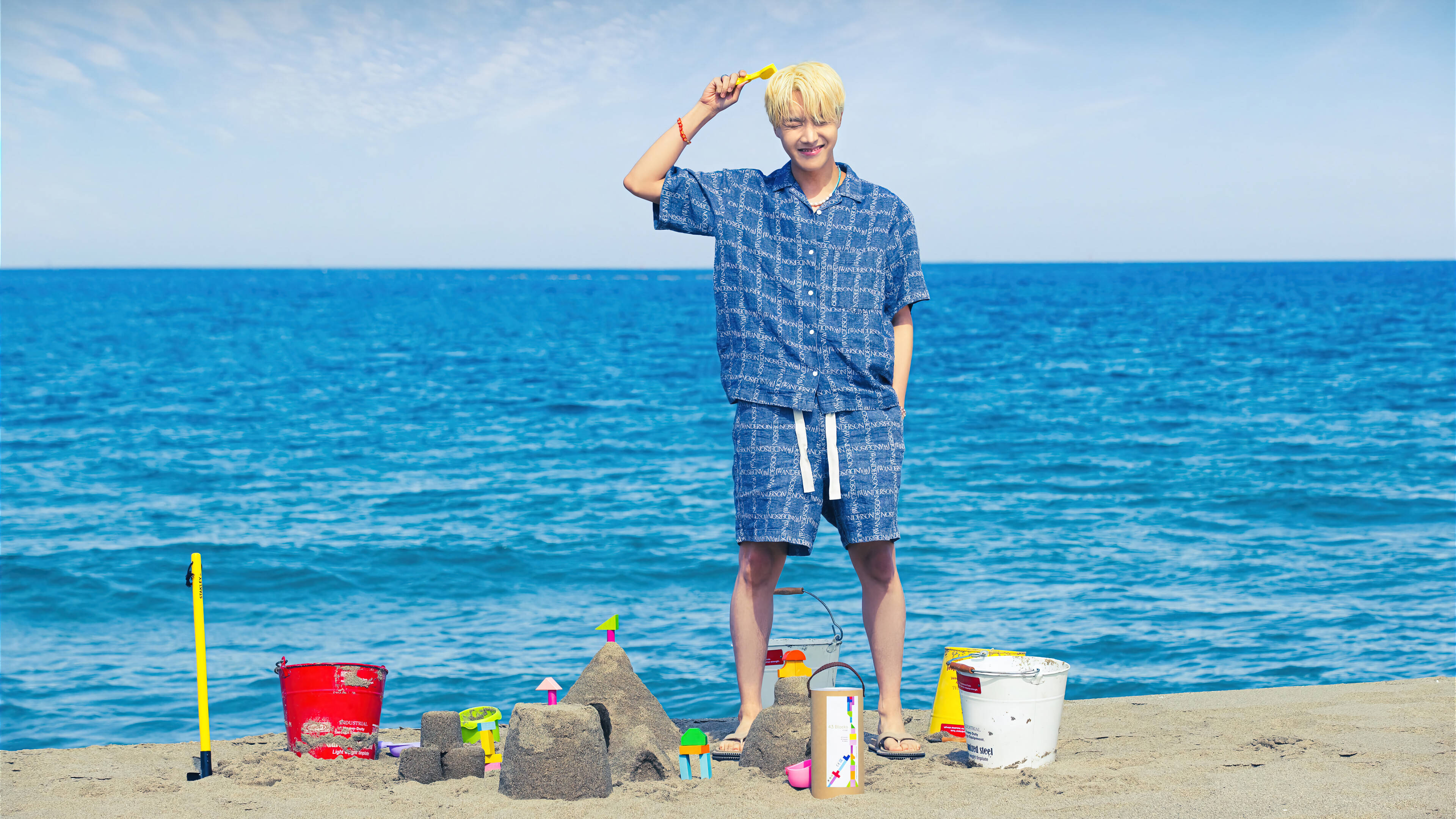 j-hope 桌布 壁紙 — 一位金髮年輕男子站在陽光明媚的海灘上，拿著黃色鏟子，旁邊有沙堡和色彩繽紛的沙灘玩具。