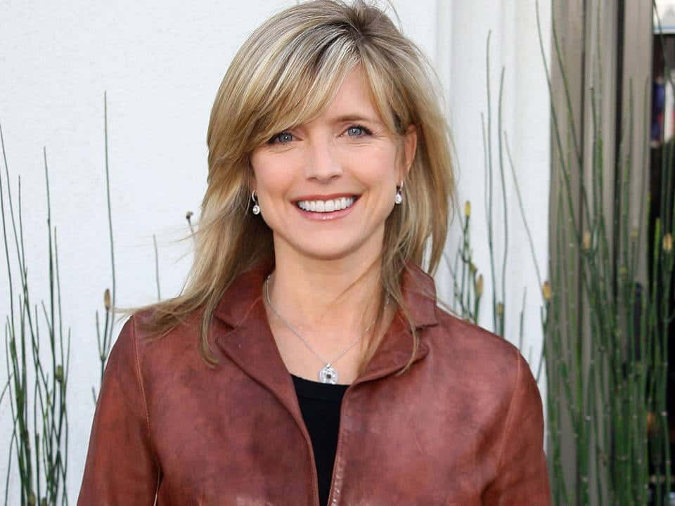 Courtney Thorne Smith 桌布 壁紙 — 一位金髮微笑的女性穿著棕色皮外套，背景是白色牆壁和綠色植物。